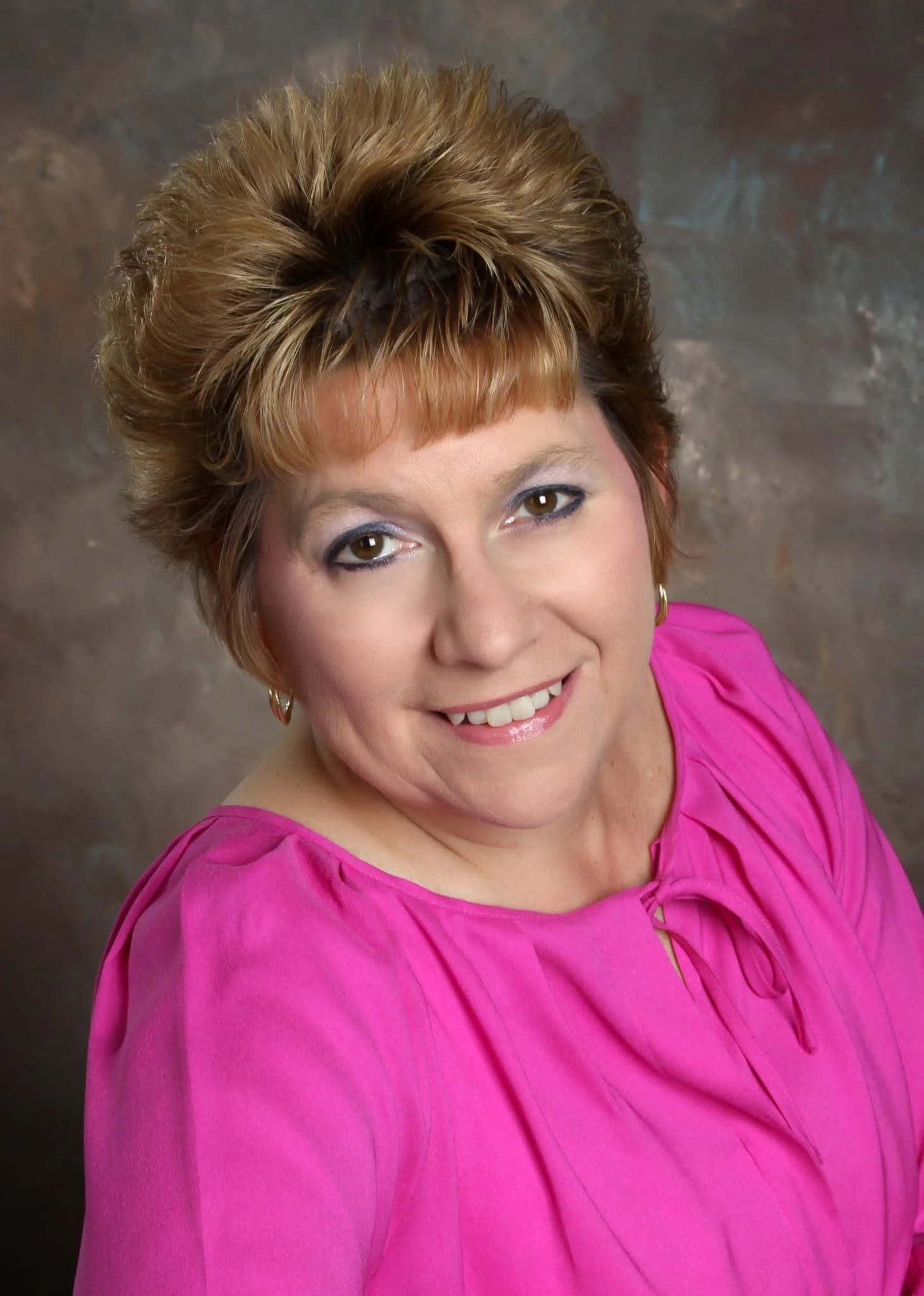 A woman with short, styled hair wearing a pink blouse and gold hoop earrings, smiling at the camera against a brown textured background.