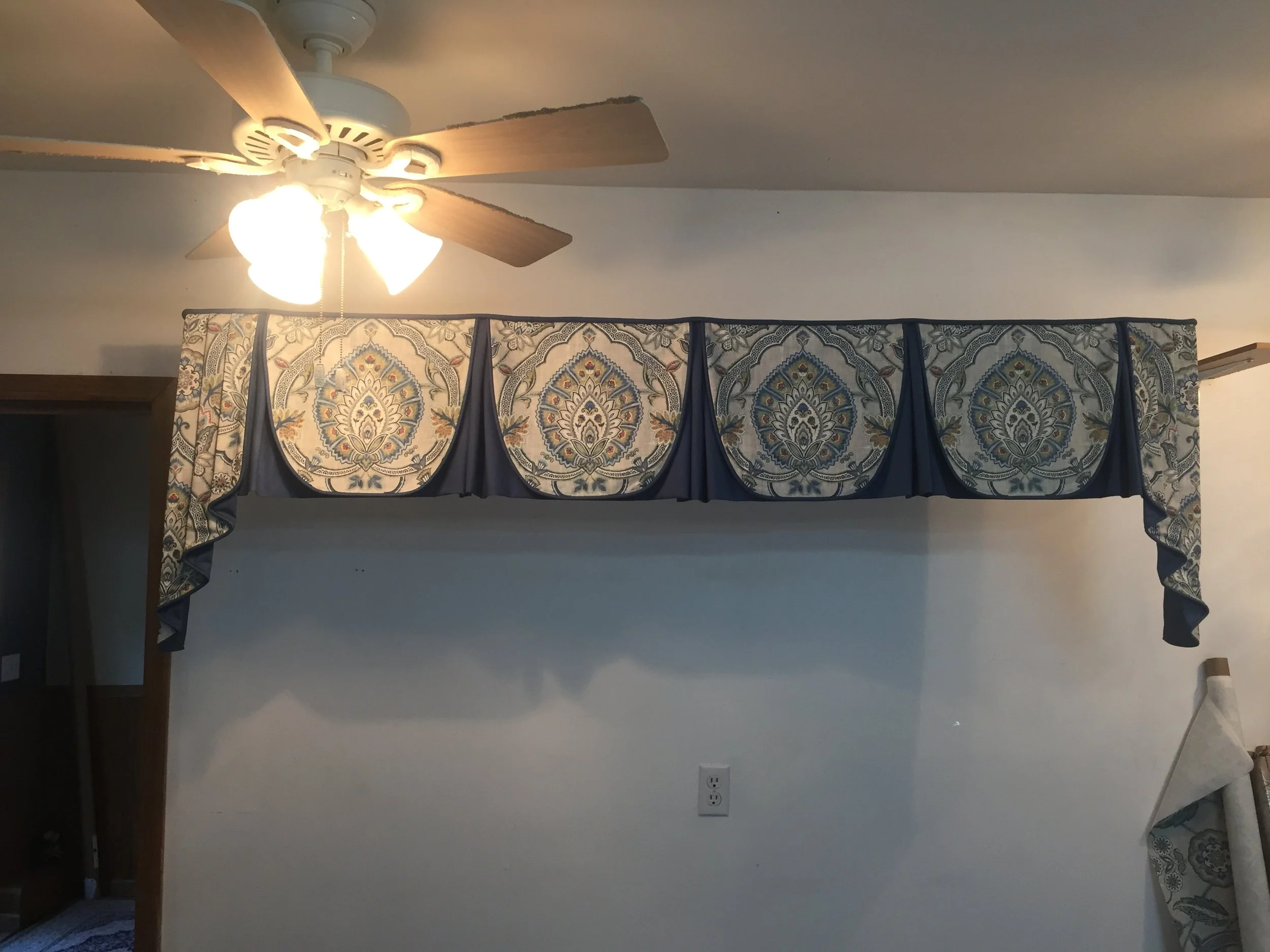 A wall-mounted valance with a blue, beige, and gold floral pattern, hanging above a blank white wall with a standard electrical outlet and a ceiling fan with four wooden blades and three light fixtures