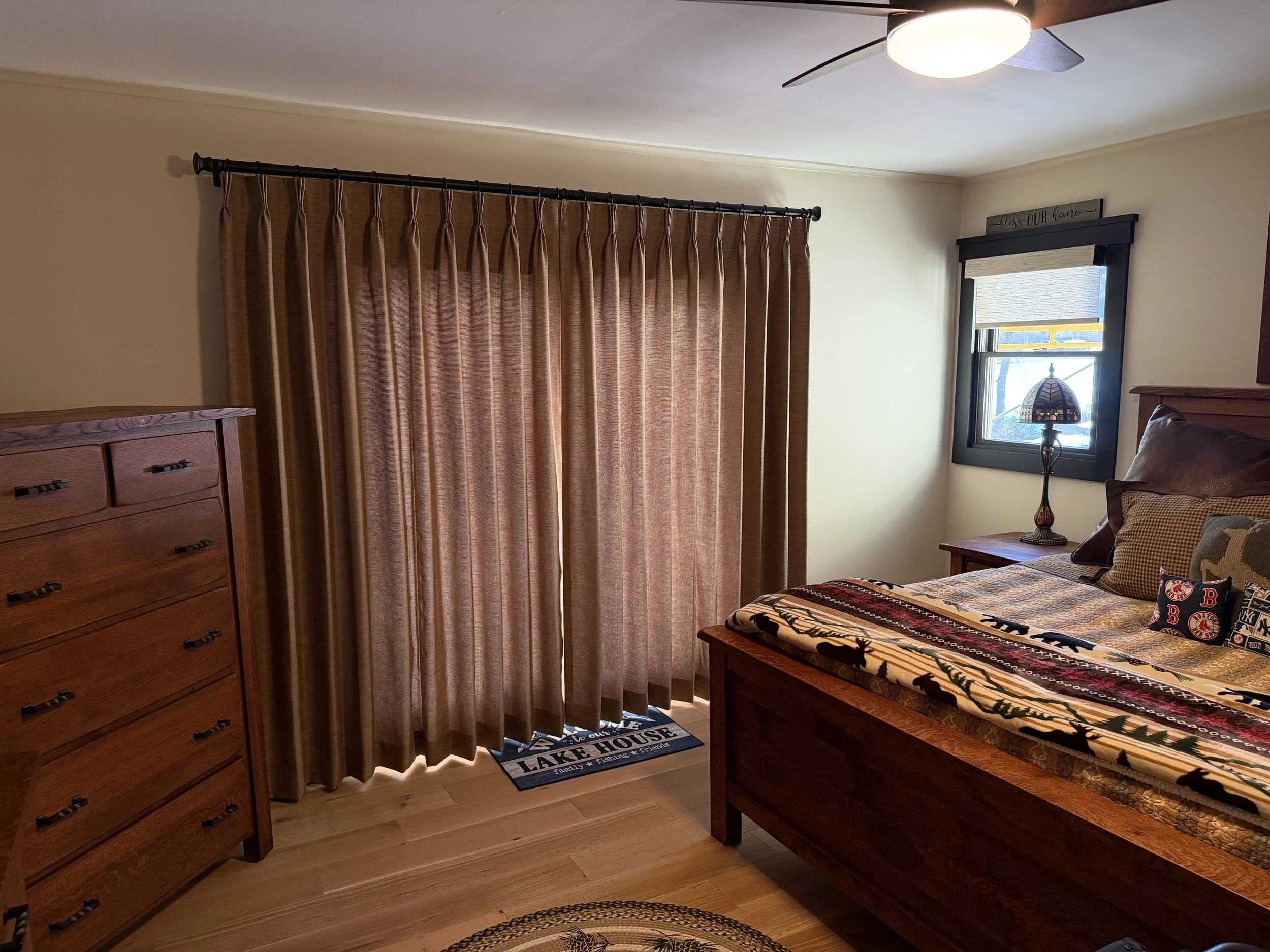 A cozy bedroom with a wooden bed, a small window with a shade, a nightstand with a decorative lamp, a dresser, and gold curtains covering a sliding door.