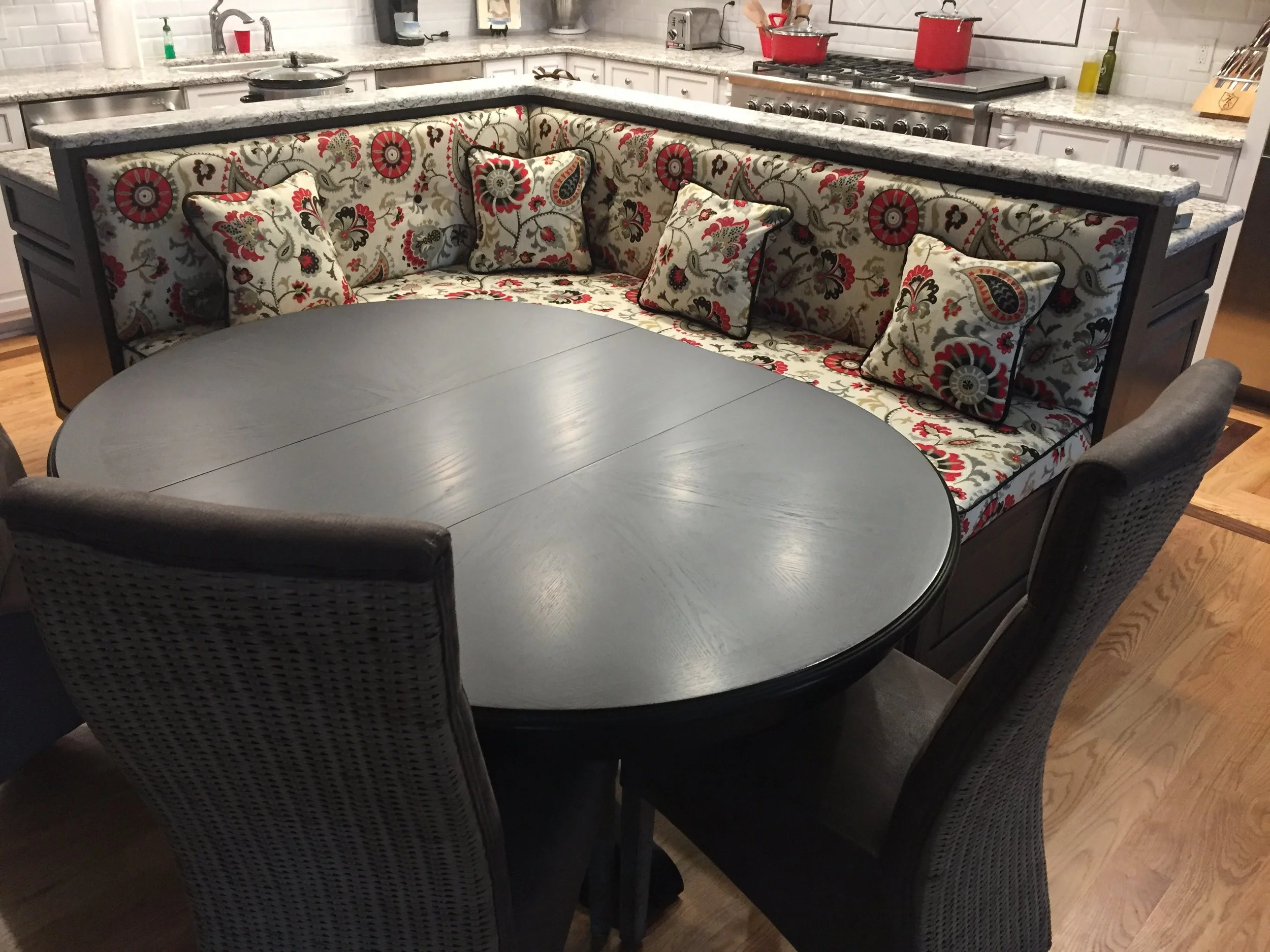 A round dining table with a gray top surrounded by four upholstered chairs and a built-in bench with floral patterned cushions and pillows, situated in a kitchen with white cabinets and granite countertops.