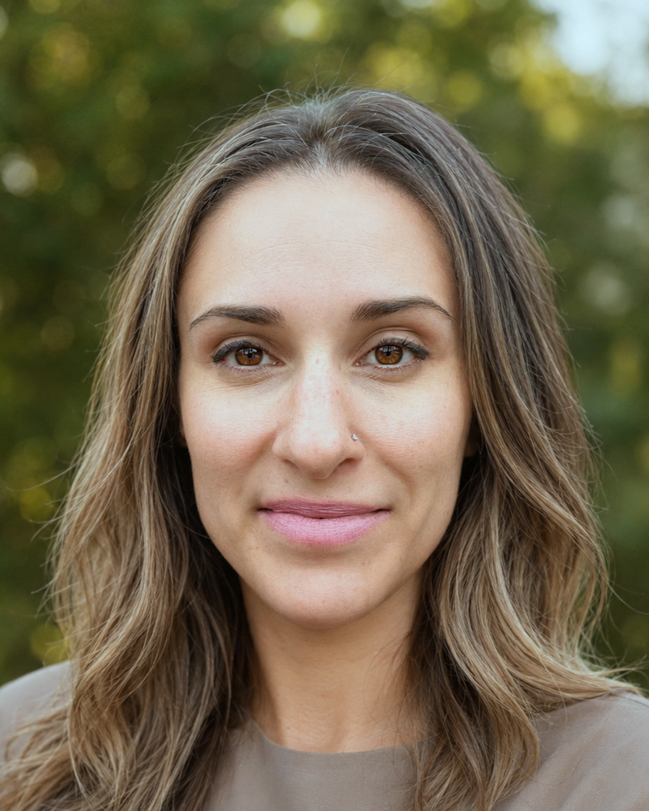 Close-up portrait of a woman with brown eyes and wavy light brown hair, outdoors with blurred green trees in the background.