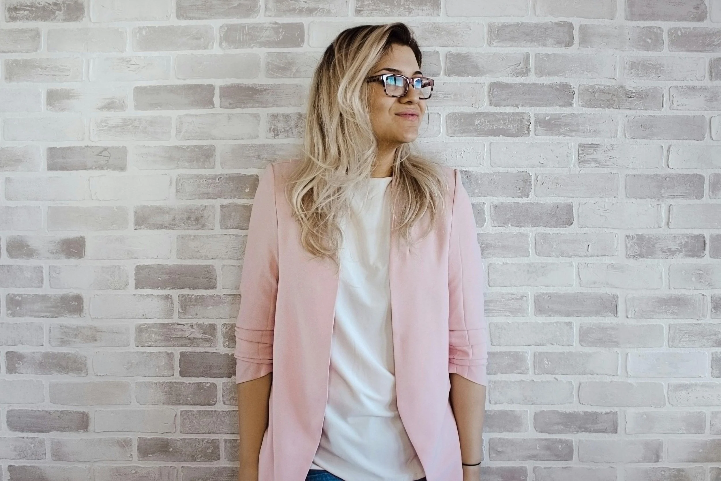 Woman with blonde hair wearing glasses, pink blazer, and white top standing against a white brick wall
