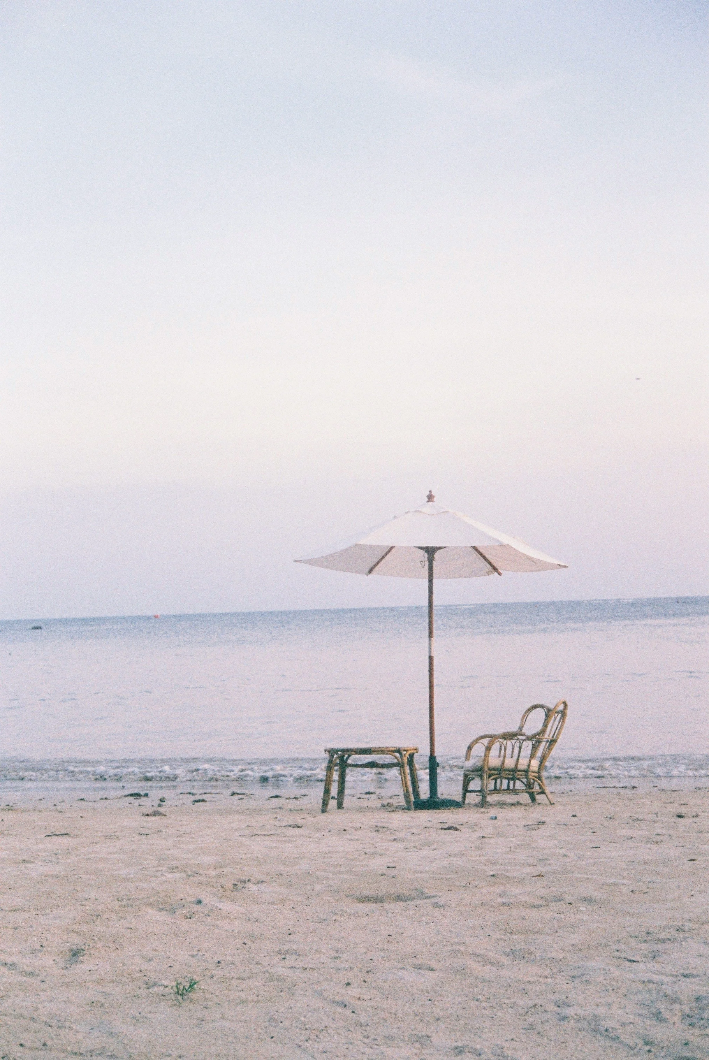 A beach scene with a large white umbrella, a couple of chairs, and a small table on sandy shore, with the ocean in the background under a cloudy sky.