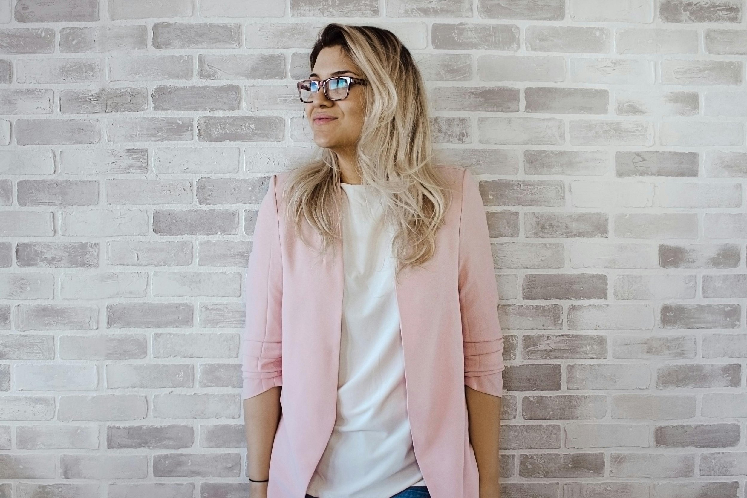 A woman with long blonde hair, wearing glasses, a light pink blazer, and a white shirt, standing against a white brick wall.