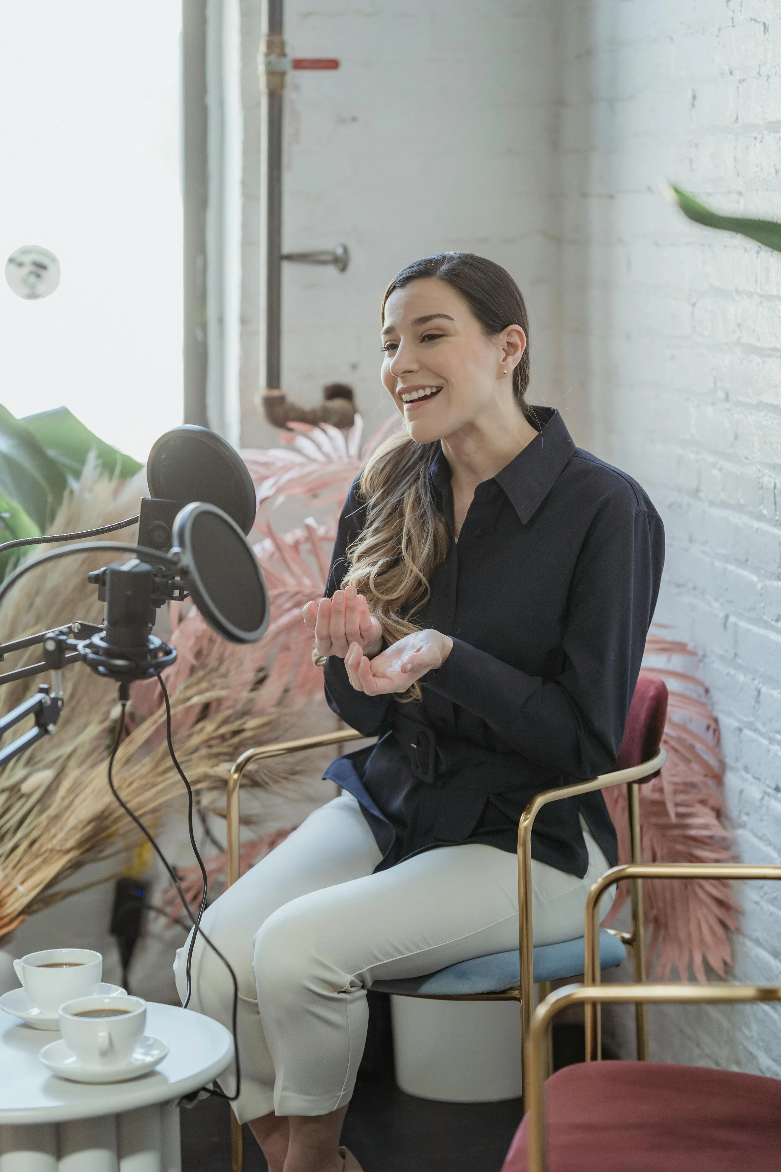 A woman seated in front of a microphone, smiling, with a white brick wall and plants in the background, and coffee cups on a small table in the foreground.