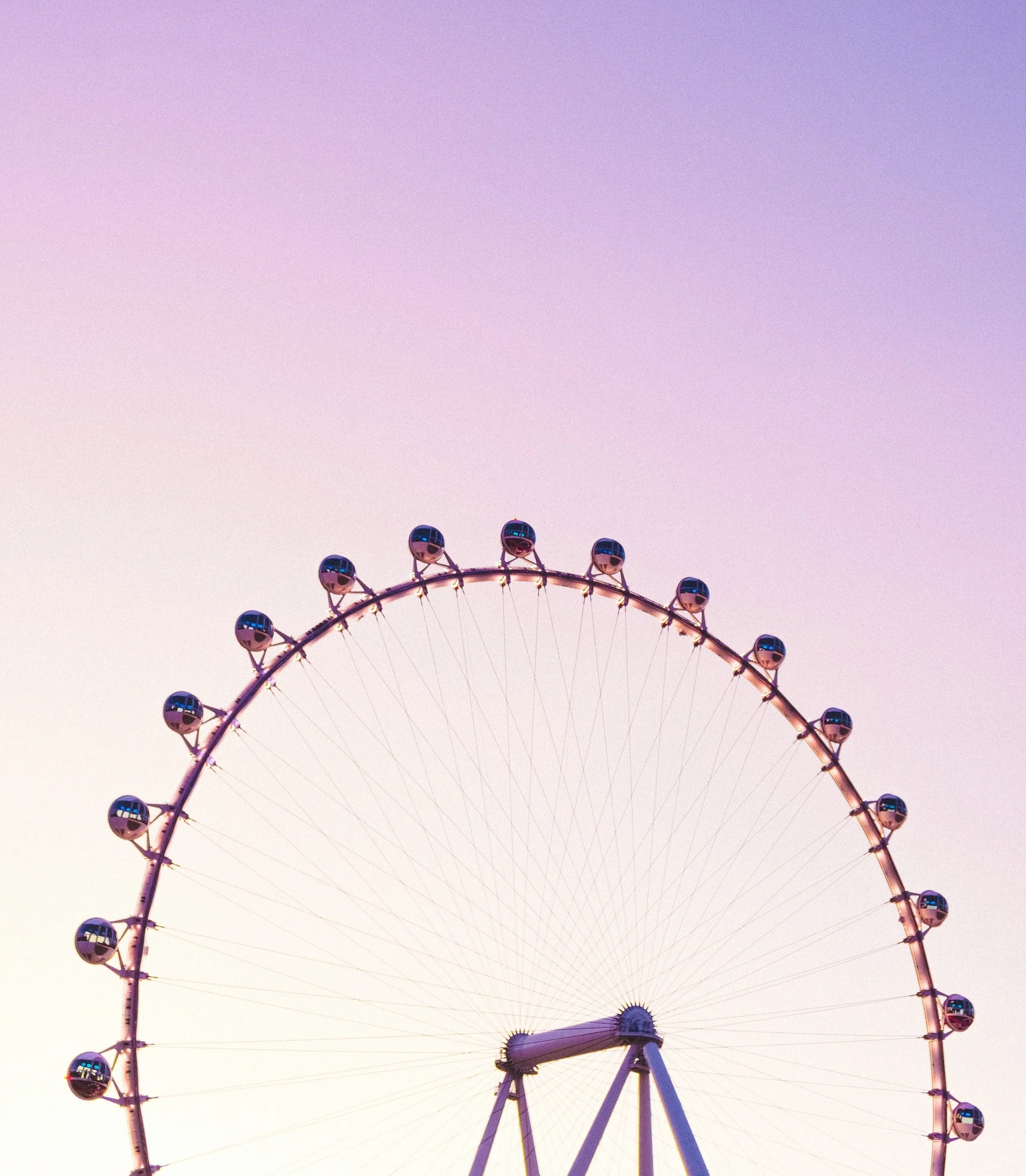 A large Ferris wheel lit up with pink and purple lights against a pastel pink and purple sky.