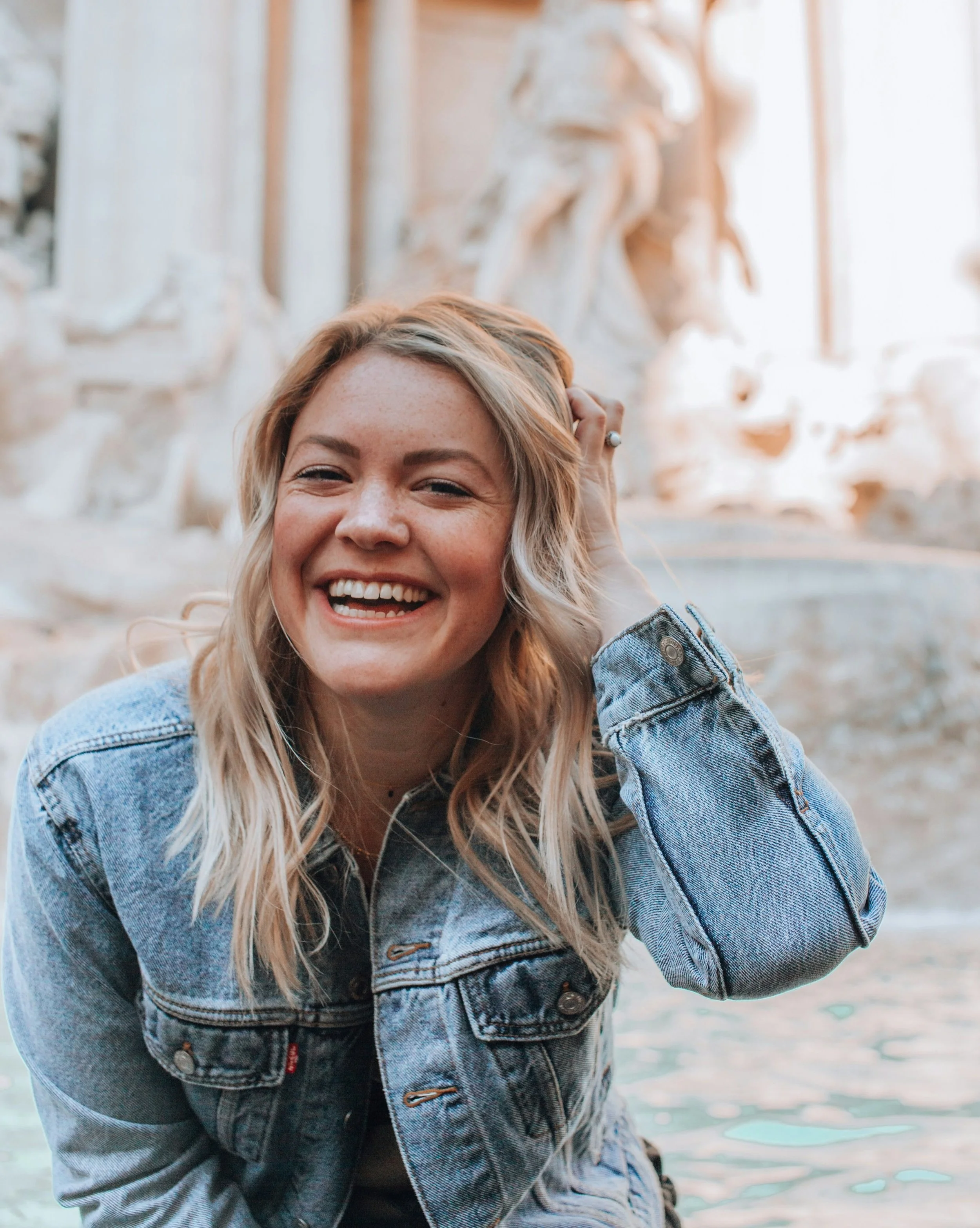 A smiling woman with blonde hair wearing a denim jacket, holding her hair back with her hand. In the background, there is a blurred fountain with water and a statue.