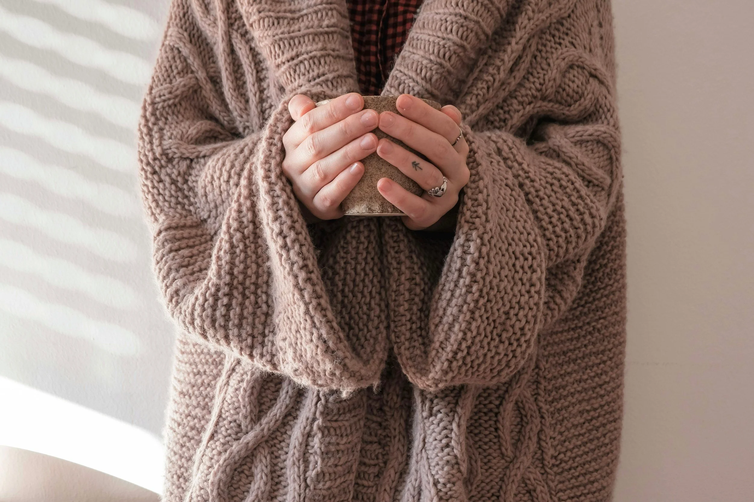 Person wearing a pink knitted cardigan holding a mug, with a white textured wall in the background.