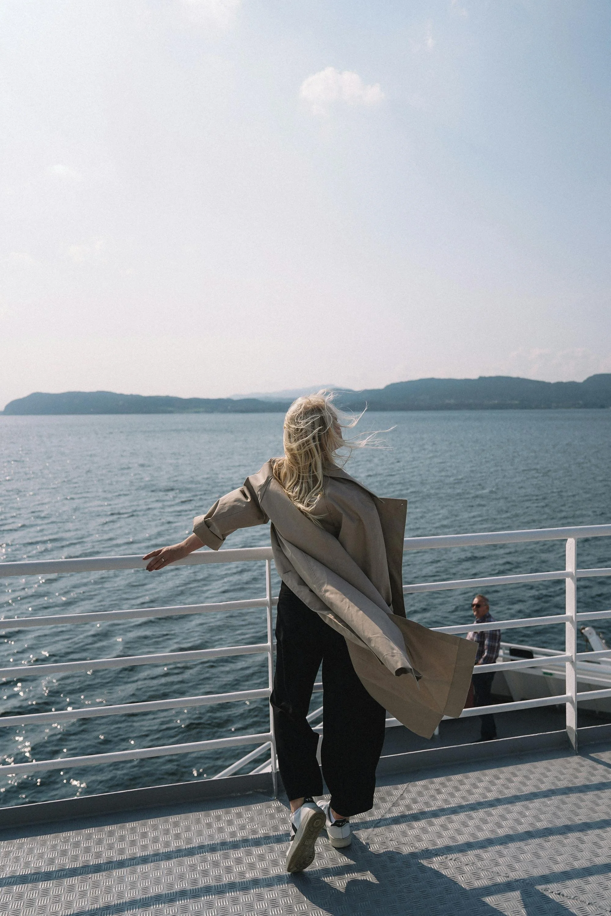 Person with blonde hair wearing a beige coat and black pants standing on the deck of a boat, holding onto the white railing, with water and distant land visible in the background.