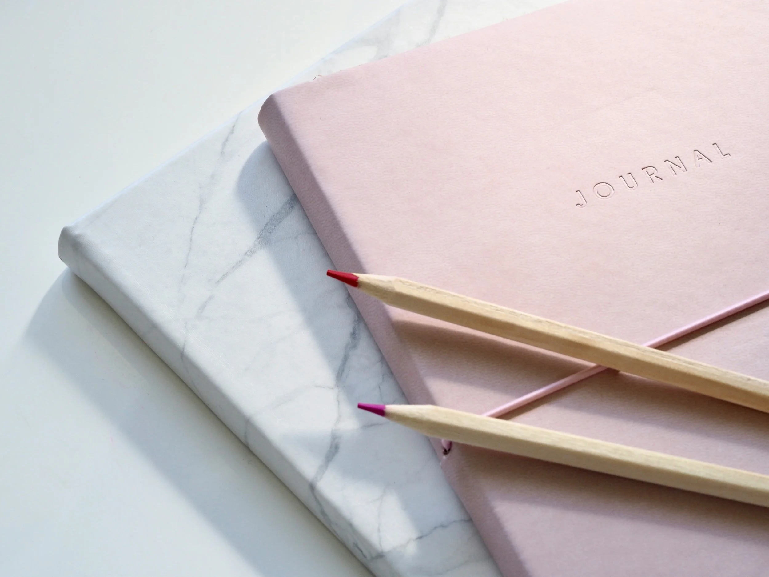 A pink journal with the word "JOURNAL" embossed on the cover, next to two colored pencils with pink and red tips, resting on a pink surface. A white marble-patterned object is partially visible underneath.