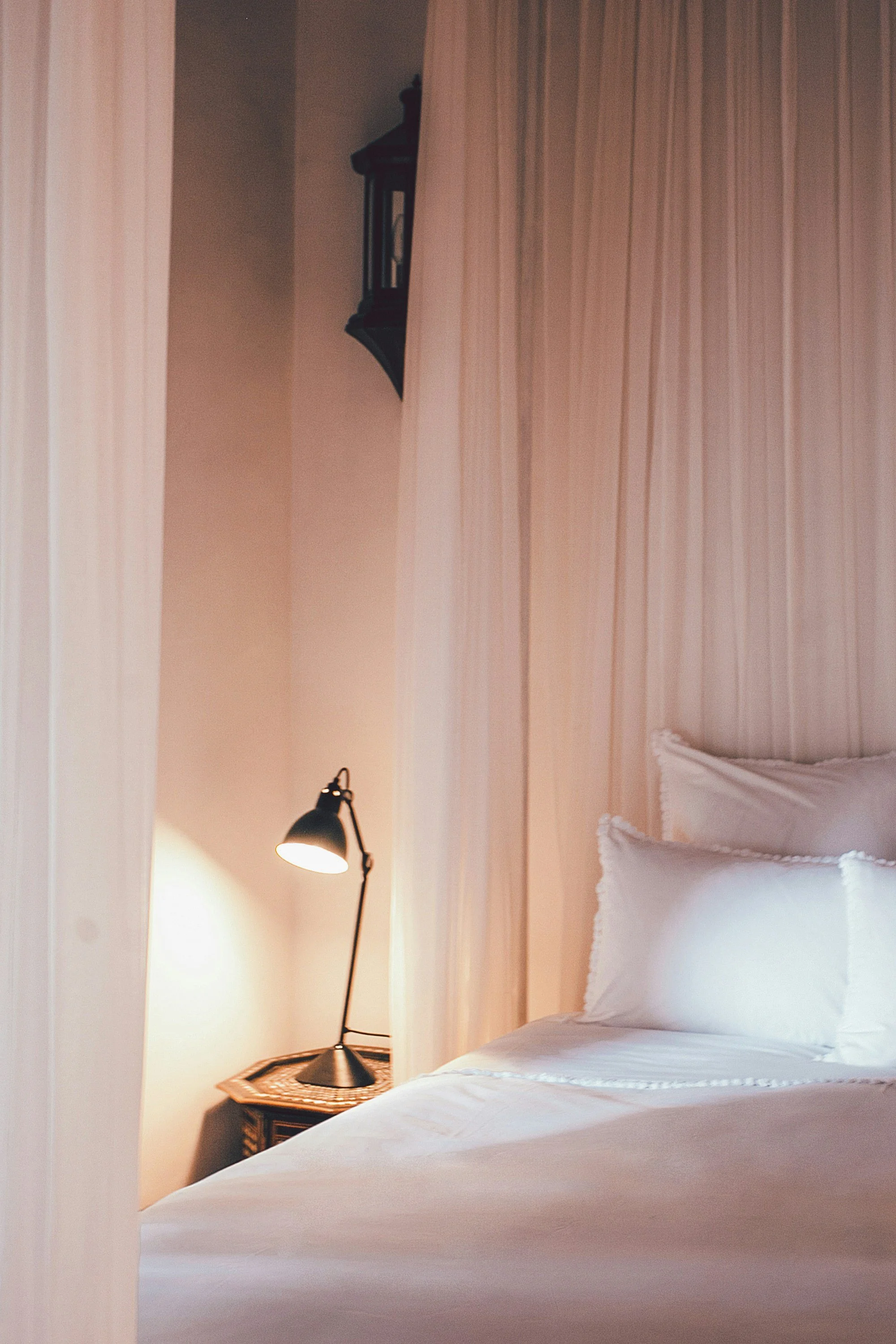 Close-up view of a neatly made bed with white sheets and pillows in a cozy bedroom. A black desk lamp is on a small wooden table beside the bed. The background features a wall with vertical beige paneling and a partially visible black wall-mounted lantern.