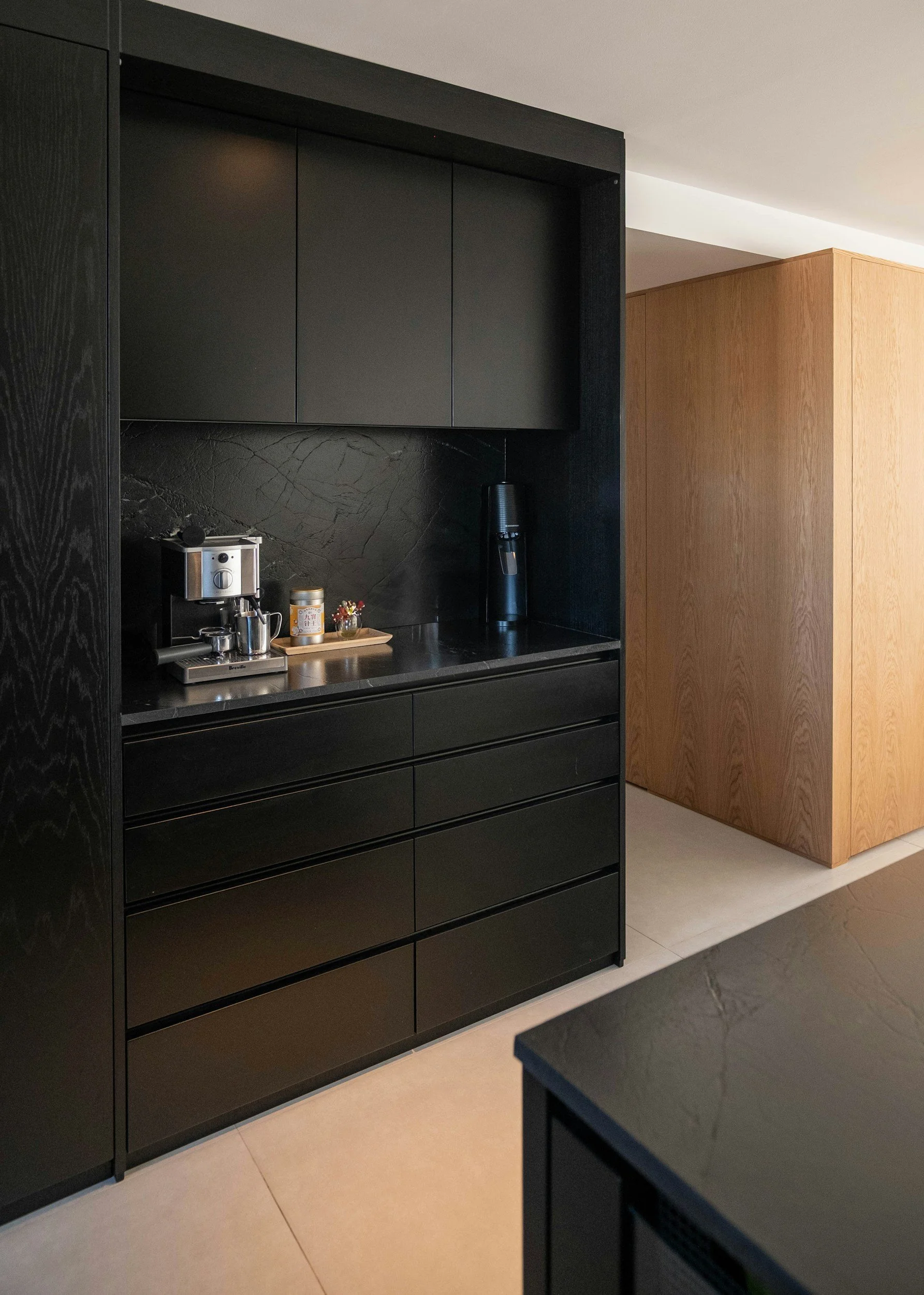 Modern kitchen area featuring black cabinetry, black marble backsplash, a coffee machine, a coffee mug, a small jar, a black electric kettle, and wooden panels.