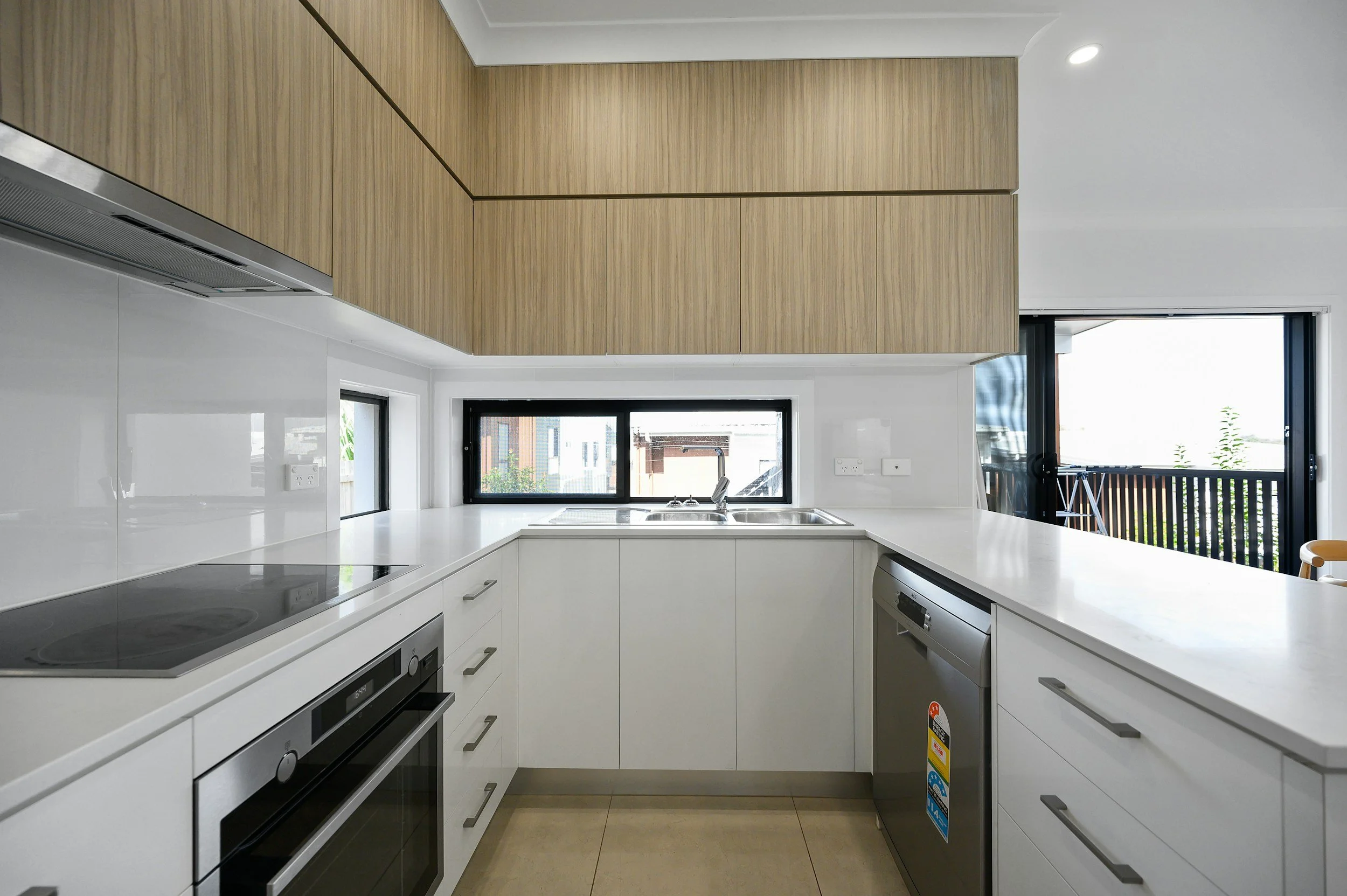 Kitchen with white cabinets, a black cooktop, a built-in oven, and a stainless steel dishwasher, with a window above the sink and a sliding door to a balcony with a railing.