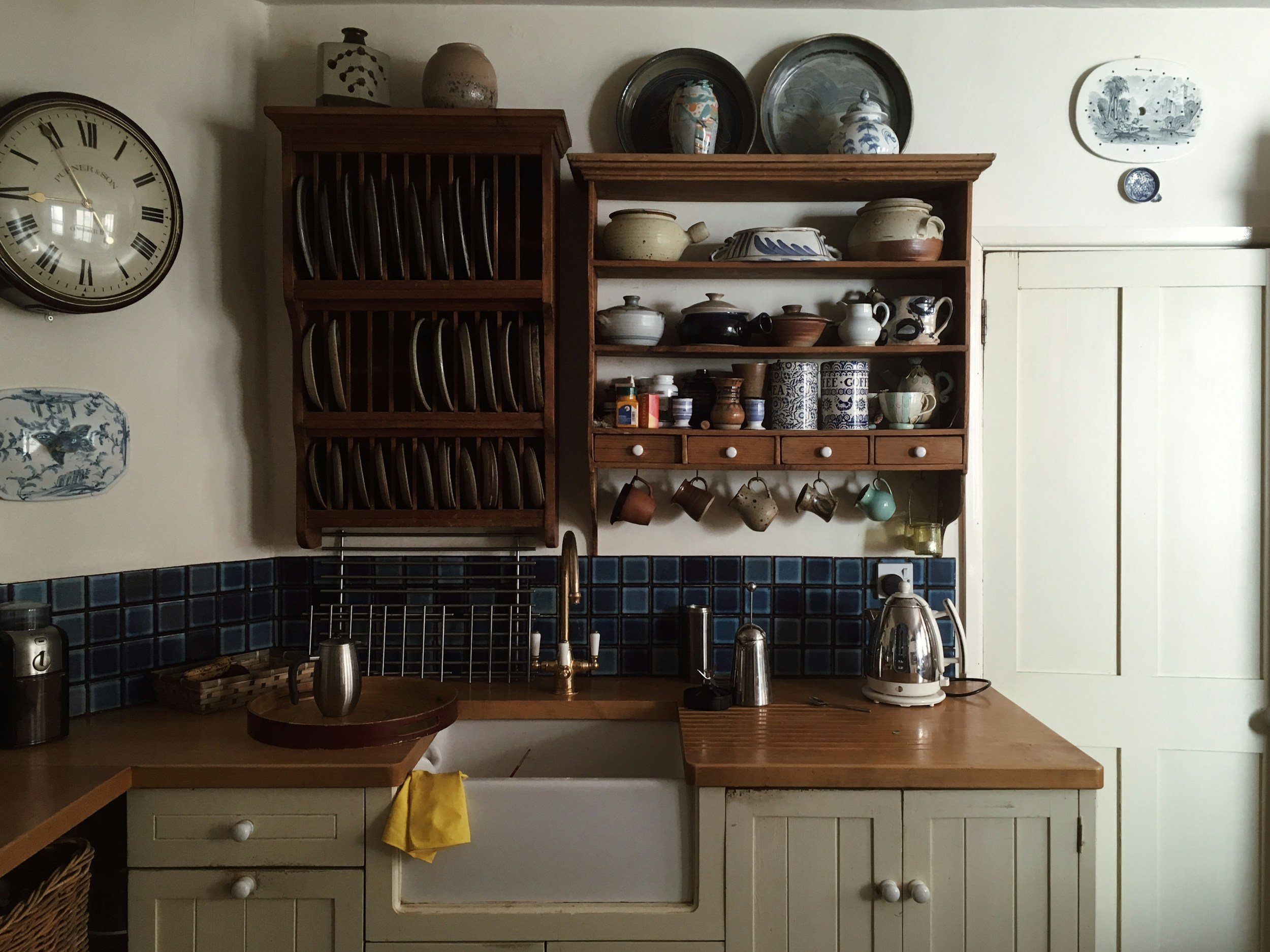 A kitchen scene with wooden shelves holding dishes, bowls, and kitchenware. There is a large wall clock showing the time as approximately 11:52. The countertops have various appliances and utensils, and the cabinetry below has white drawers and doors.