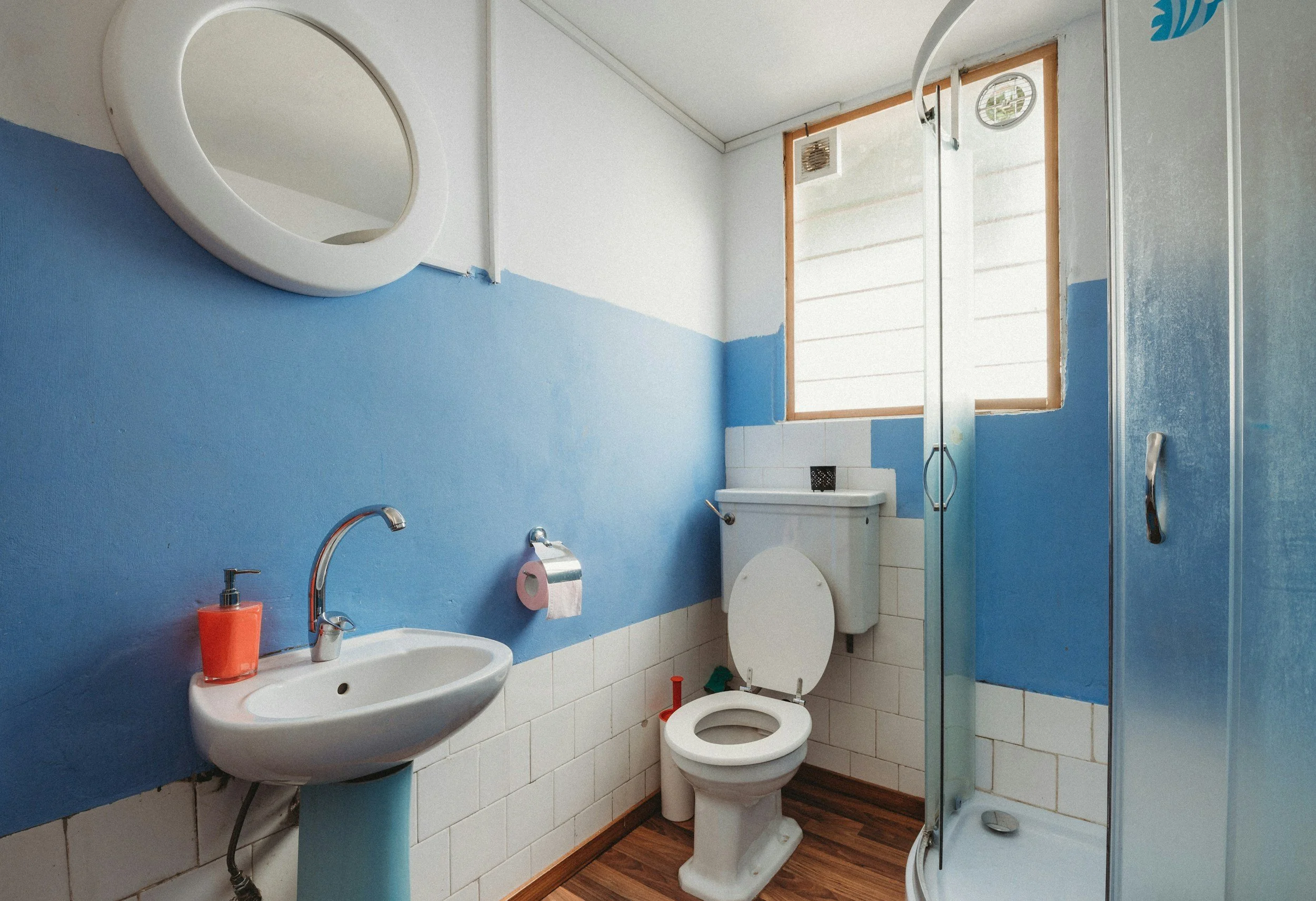 Bathroom with white and blue walls, a mirror, a sink with an orange soap dispenser, a toilet, and a shower stall. There's a window above the toilet.