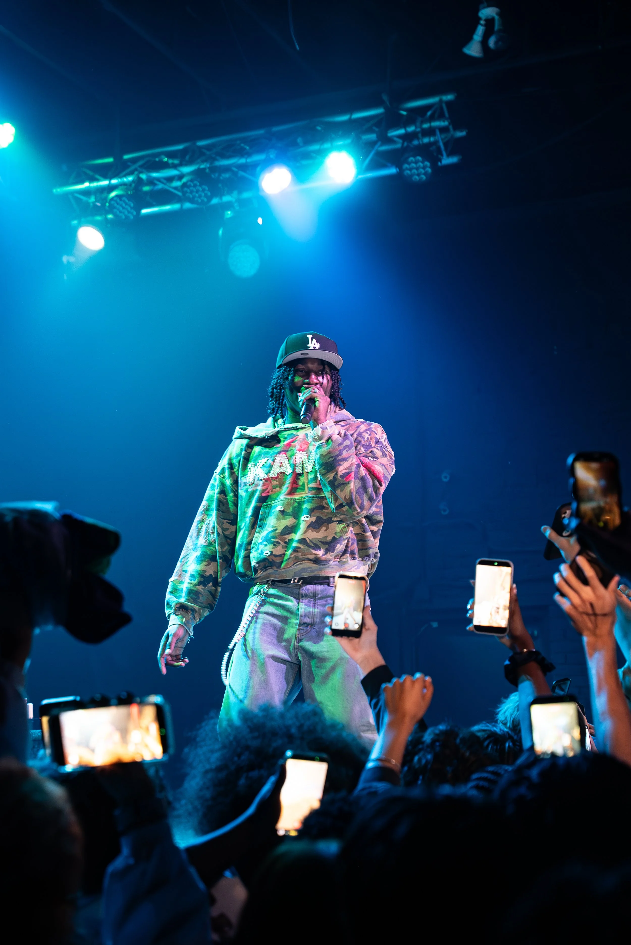 Performer on stage singing into a microphone at a concert, wearing a camouflage hoodie, light-colored jeans, and a black cap with white letters, with blue and purple stage lights overhead and audience members taking photos.