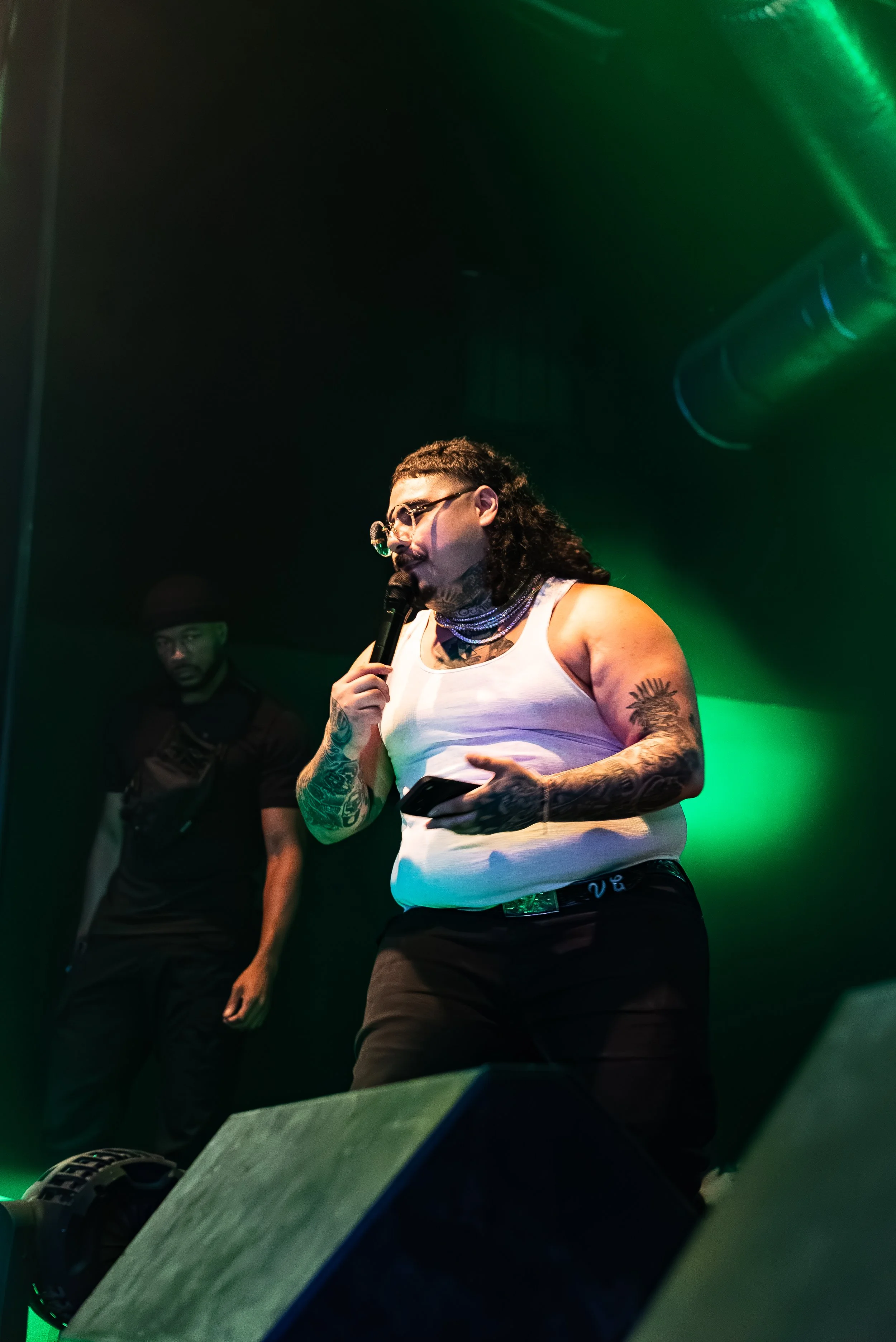 That Mexican OT wearing glasses, a white tank top, and jewelry holding a microphone and a phone on stage with green lighting, with another person standing in the background.