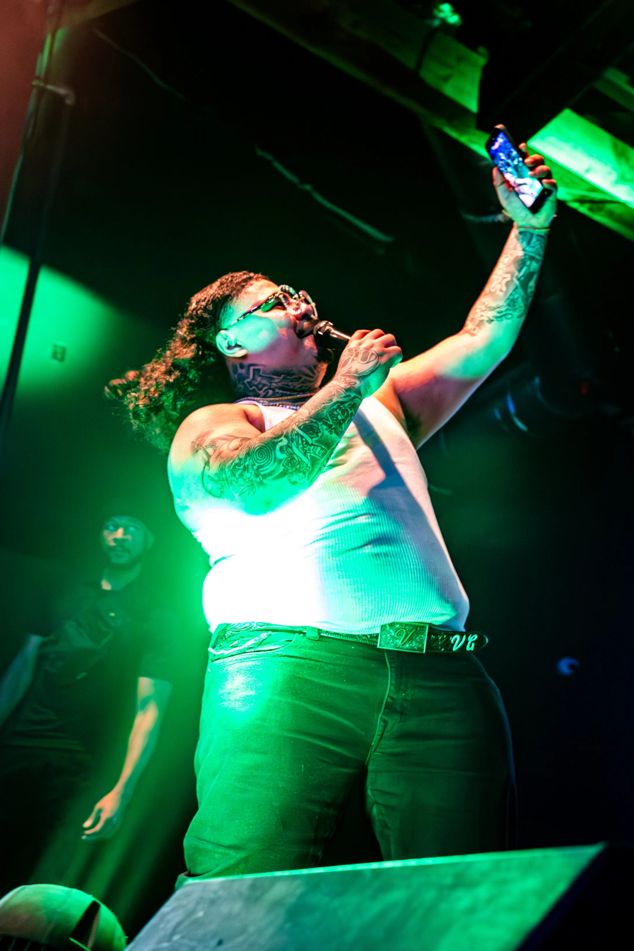 That Mexican OT, wearing glasses, a tank top, and dark pants, holding a microphone in one hand and a fans smartphone in the other, singing or speaking at a concert or live event, with green lighting and a person in the background.