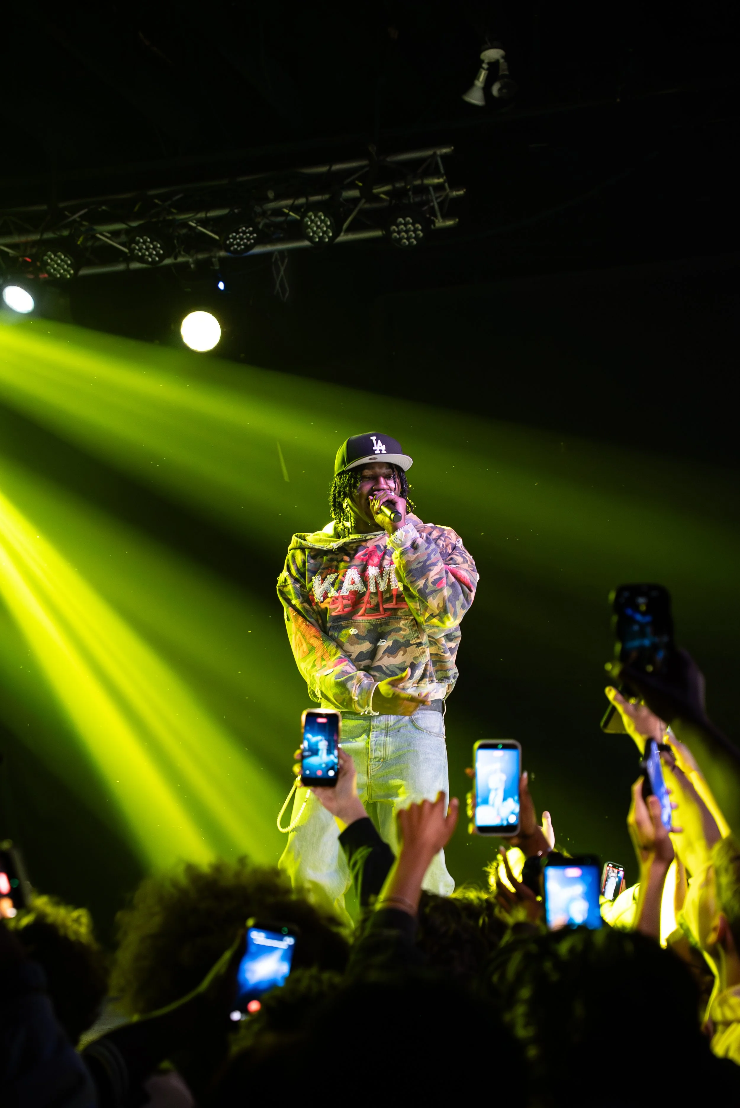Nino Paid on stage with yellow spotlight, wearing a camouflage jacket and LA baseball cap, holding microphone, audience recording with phones.