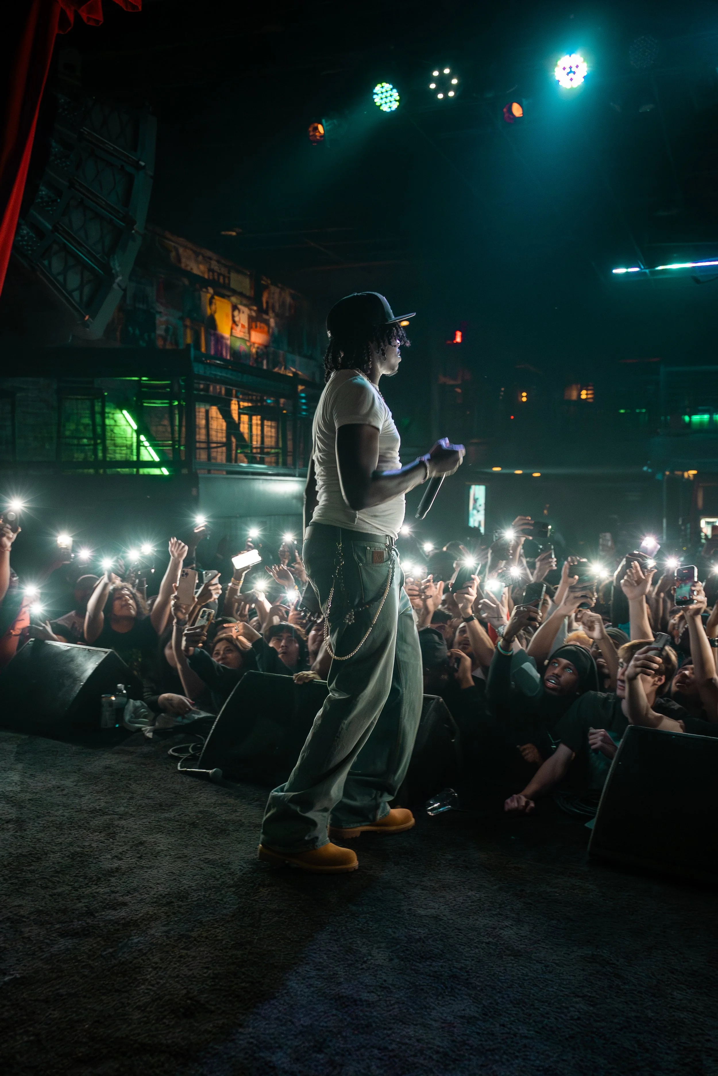 Performer on stage at a concert holding a microphone, with an audience capturing the moment with their phones, lit by stage lights.