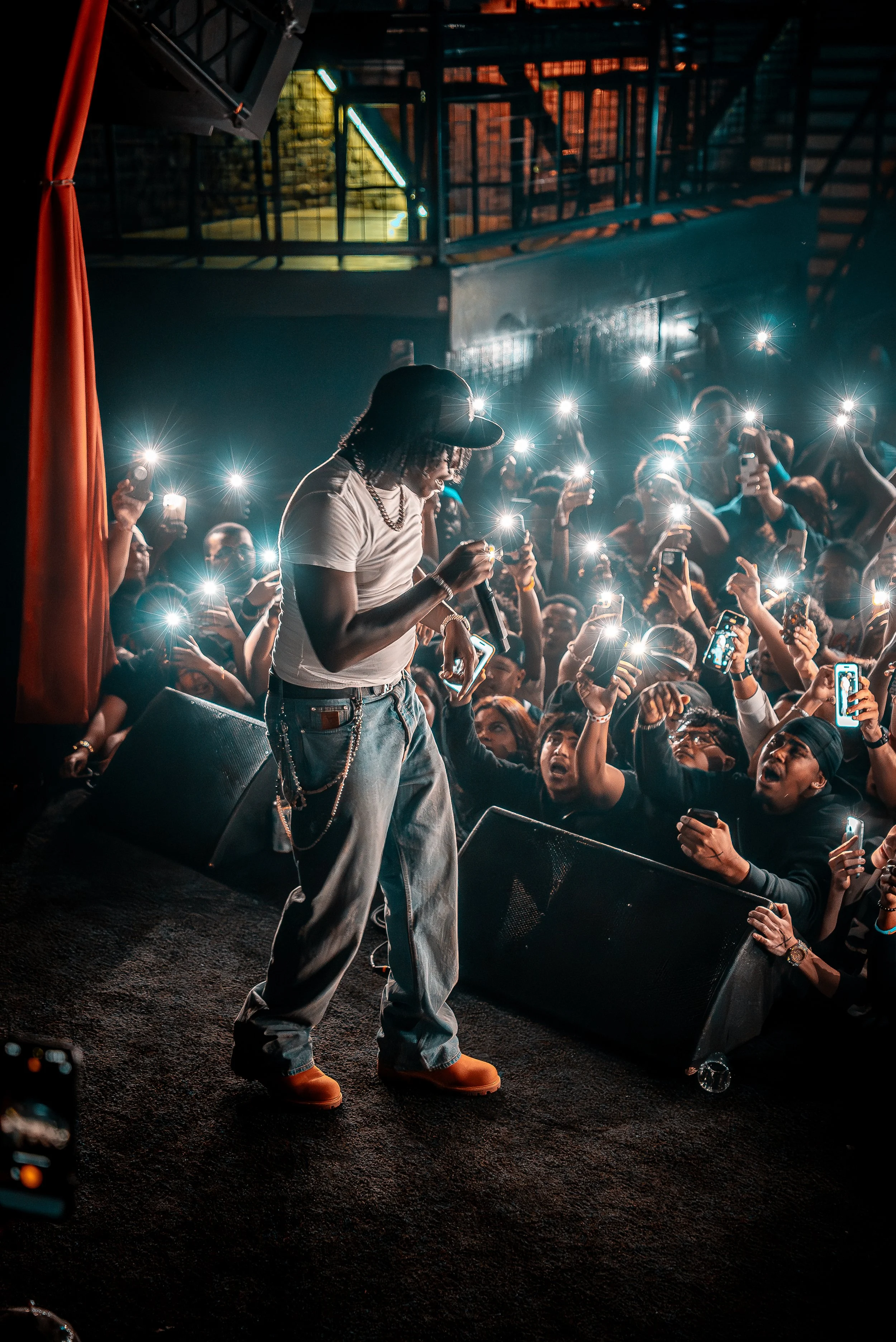 A performer on stage at a concert, wearing a black cap, white t-shirt, baggy jeans, and orange shoes, singing into a microphone. The crowd is holding up phones with flashlights on, illuminating the scene.