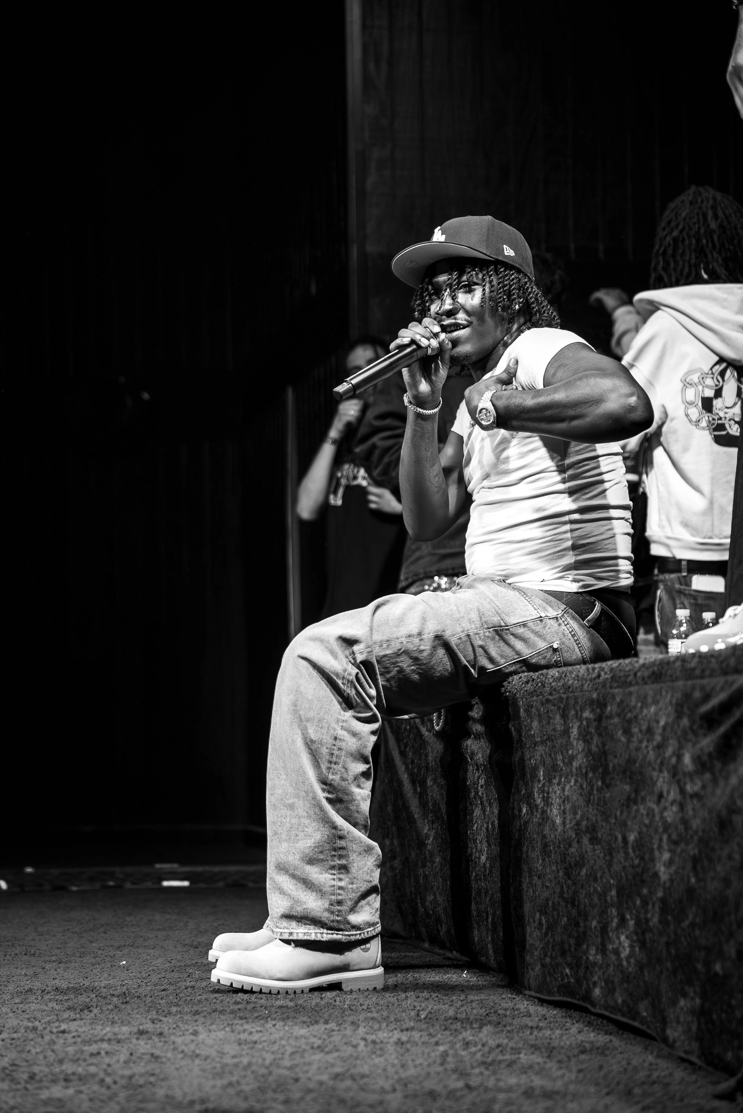 A man wearing a baseball cap and casual clothes, sitting on a stage, singing into a microphone. He has dreadlocks, a watch, and rings, with a dark background and other people standing nearby.
