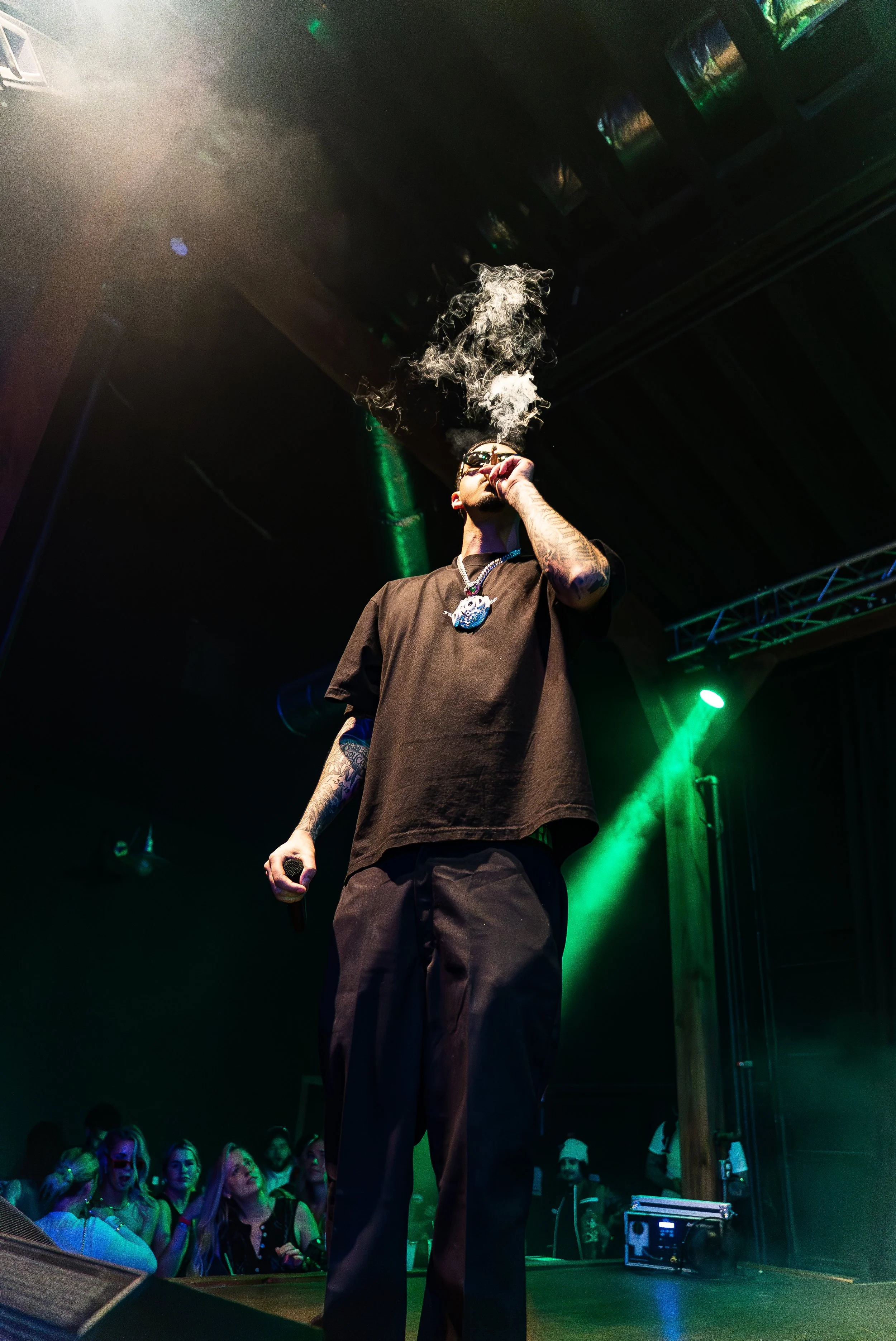 Drodi smoking on stage during a concert with audience in background, green stage lighting, and fog.