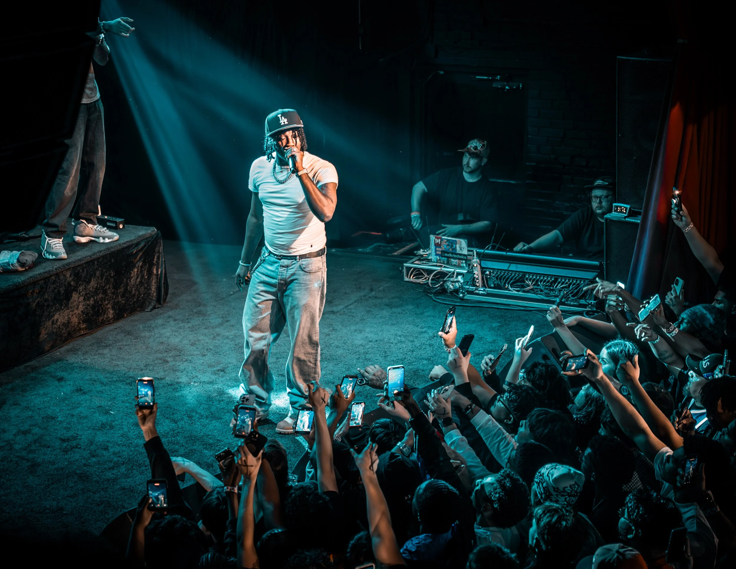 A performer on stage with a microphone, wearing a white T-shirt, jeans, a black LA cap, and jewelry, captivating a crowd in a dimly lit venue. Fans in the audience are holding up phones, recording or taking photos of the performance.
