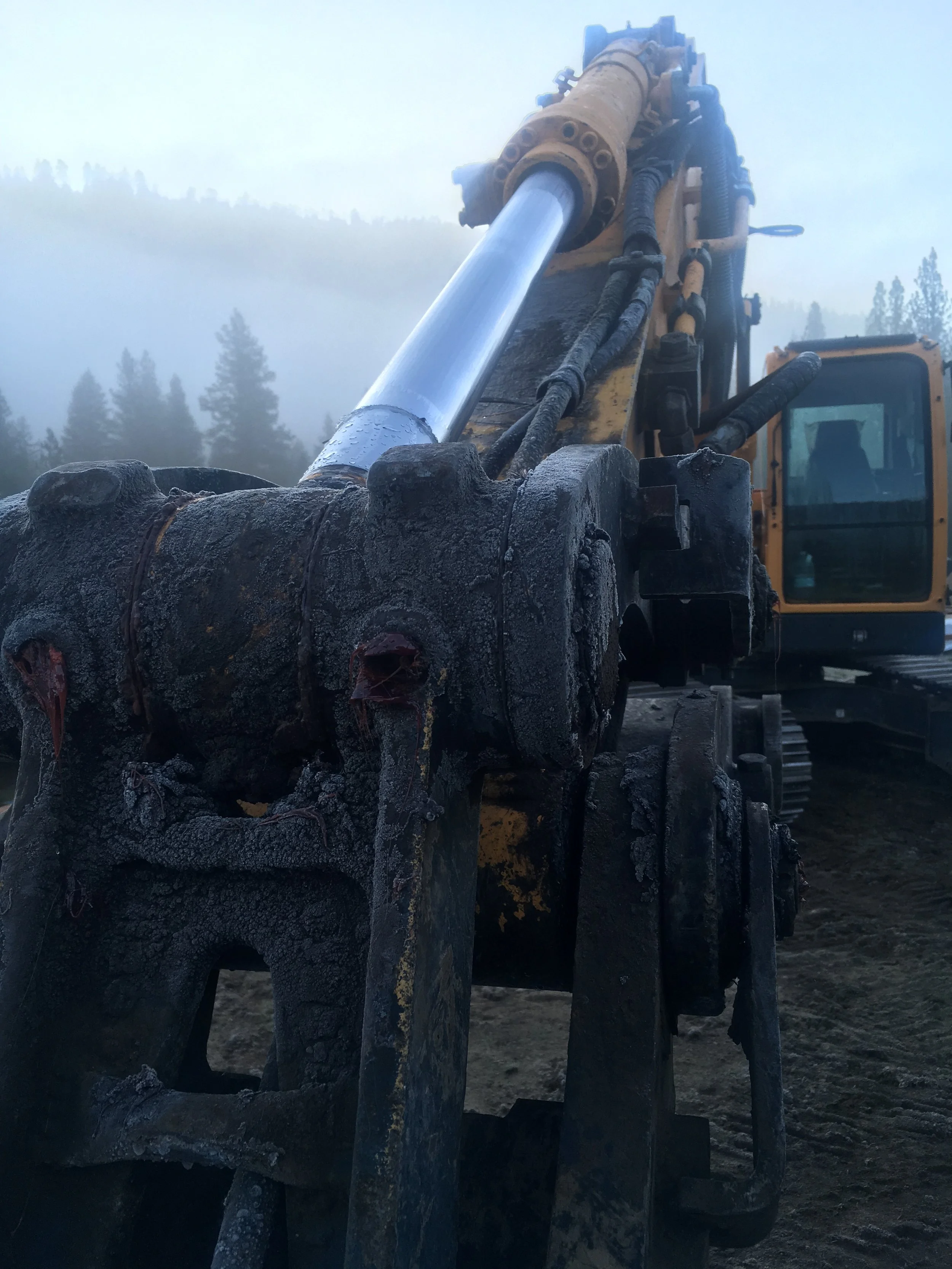 Close-up of a large hydraulic excavator arm with a dirty, blackened bucket at a construction site, with misty forested hills in the background.