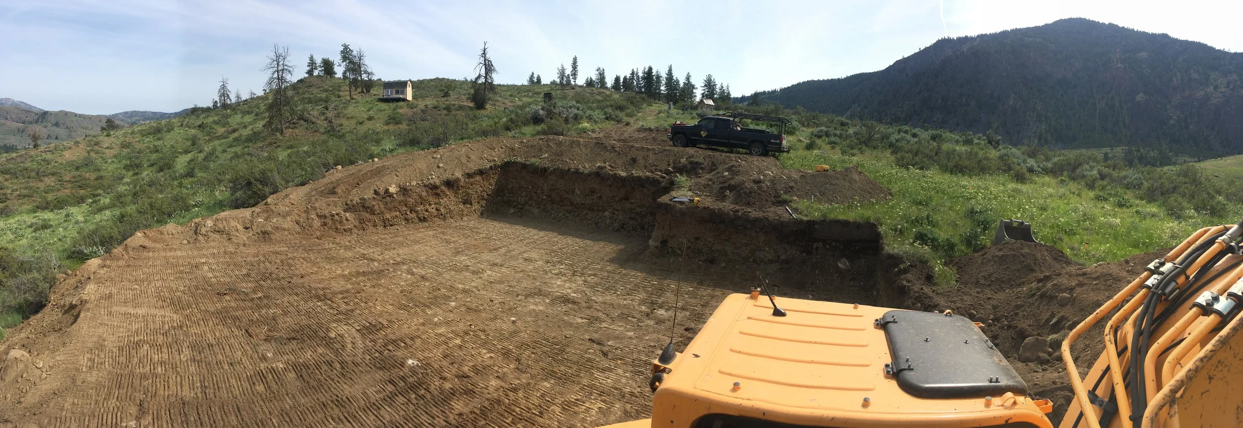 Construction site with bulldozer preparing land, hillside with trees, a truck, and mountains in the background