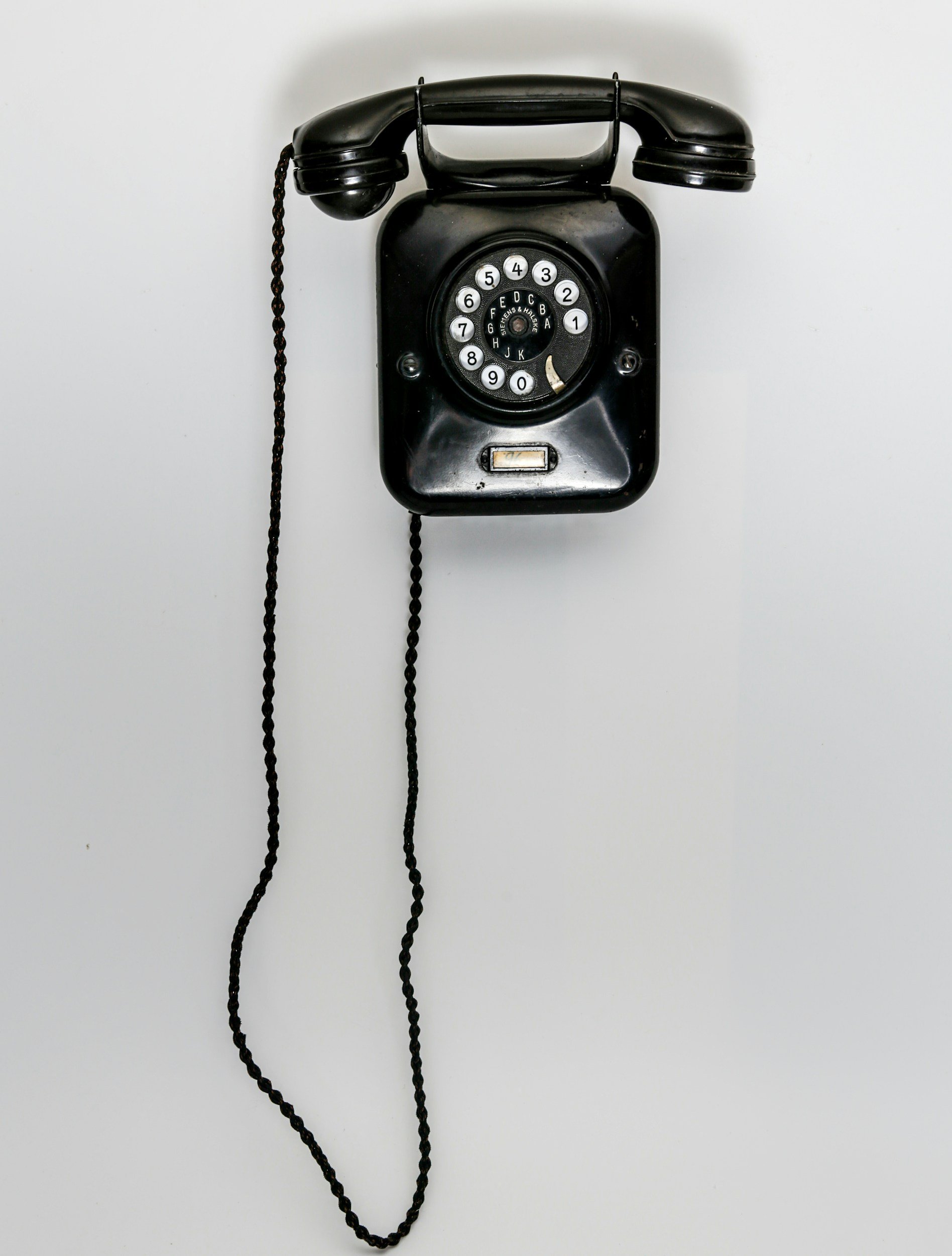 Vintage black rotary wall phone with a coiled cord hanging on a plain white wall.