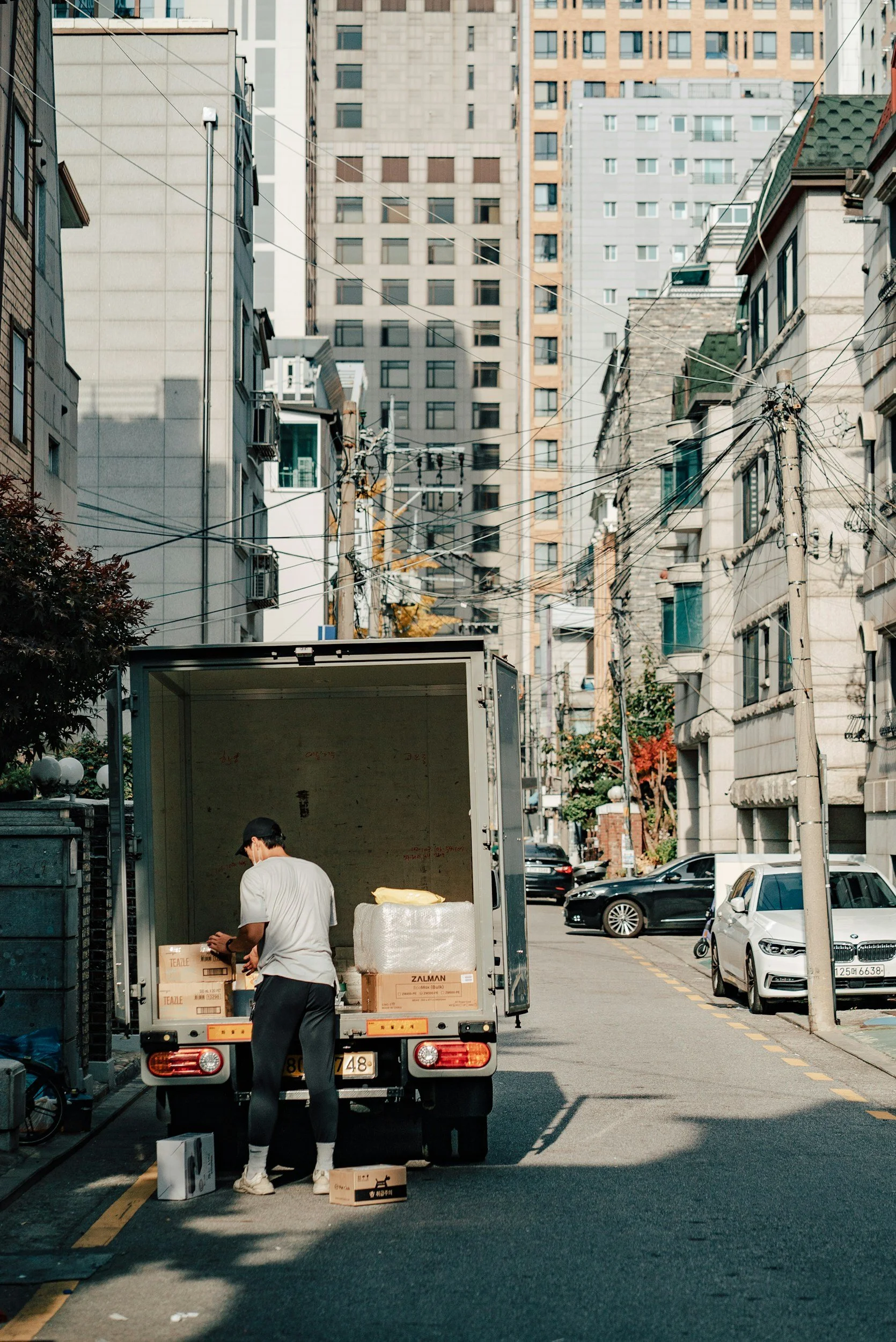 A man unloading boxes from a delivery truck on a city street, with tall buildings and parked cars in the background.