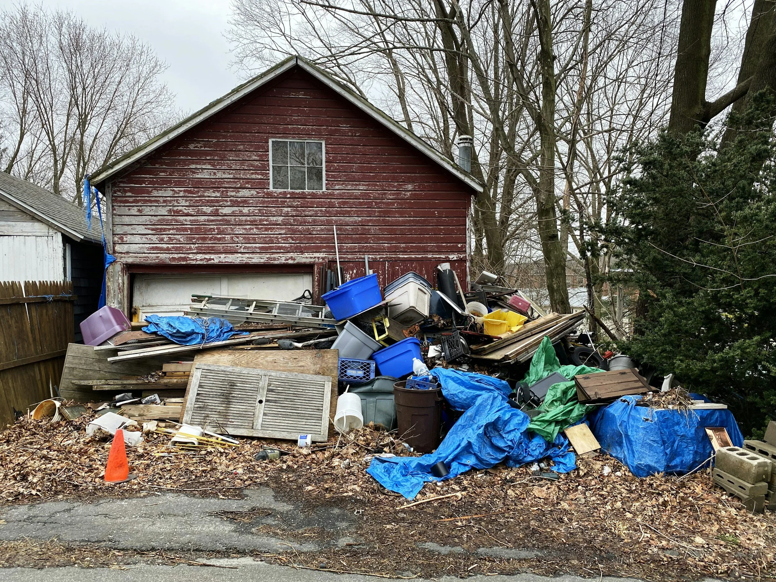 A pile of discarded items including plastic bins, wooden planks, and other debris in front of a weathered red barn.