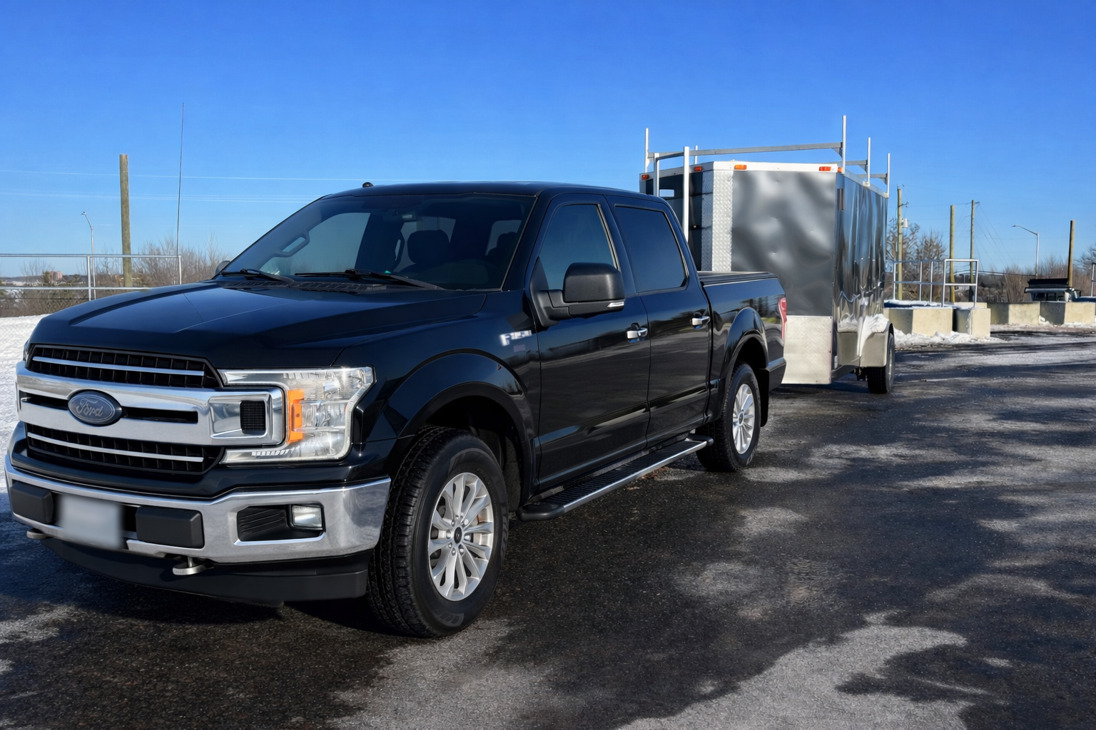 Black Ford pickup truck towing a gray enclosed trailer on a paved lot under a clear blue sky.