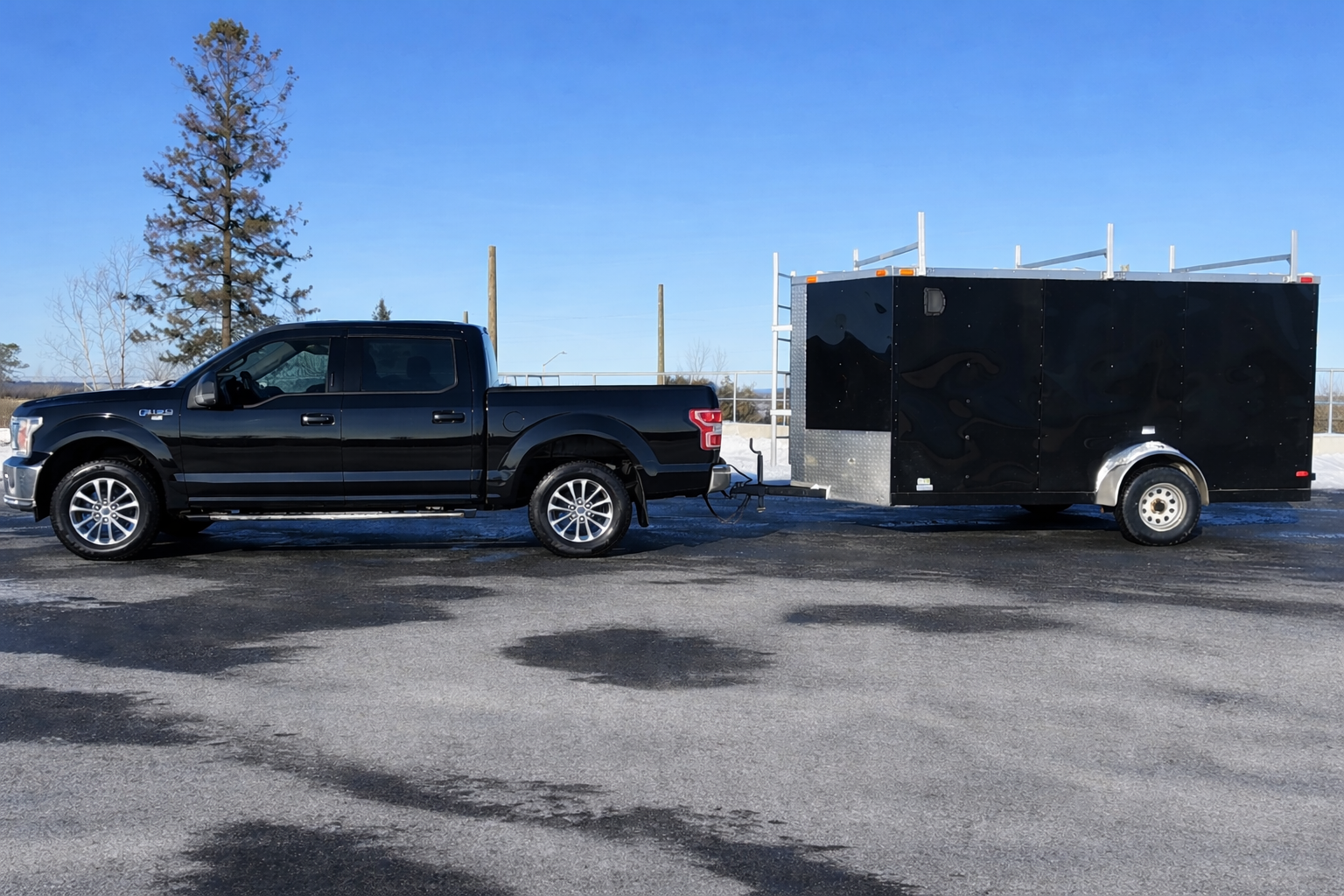 Black pickup truck at Ottawa Dump towing a large black enclosed utility trailer with a metal front panel, parked on an asphalt lot under a clear blue sky.