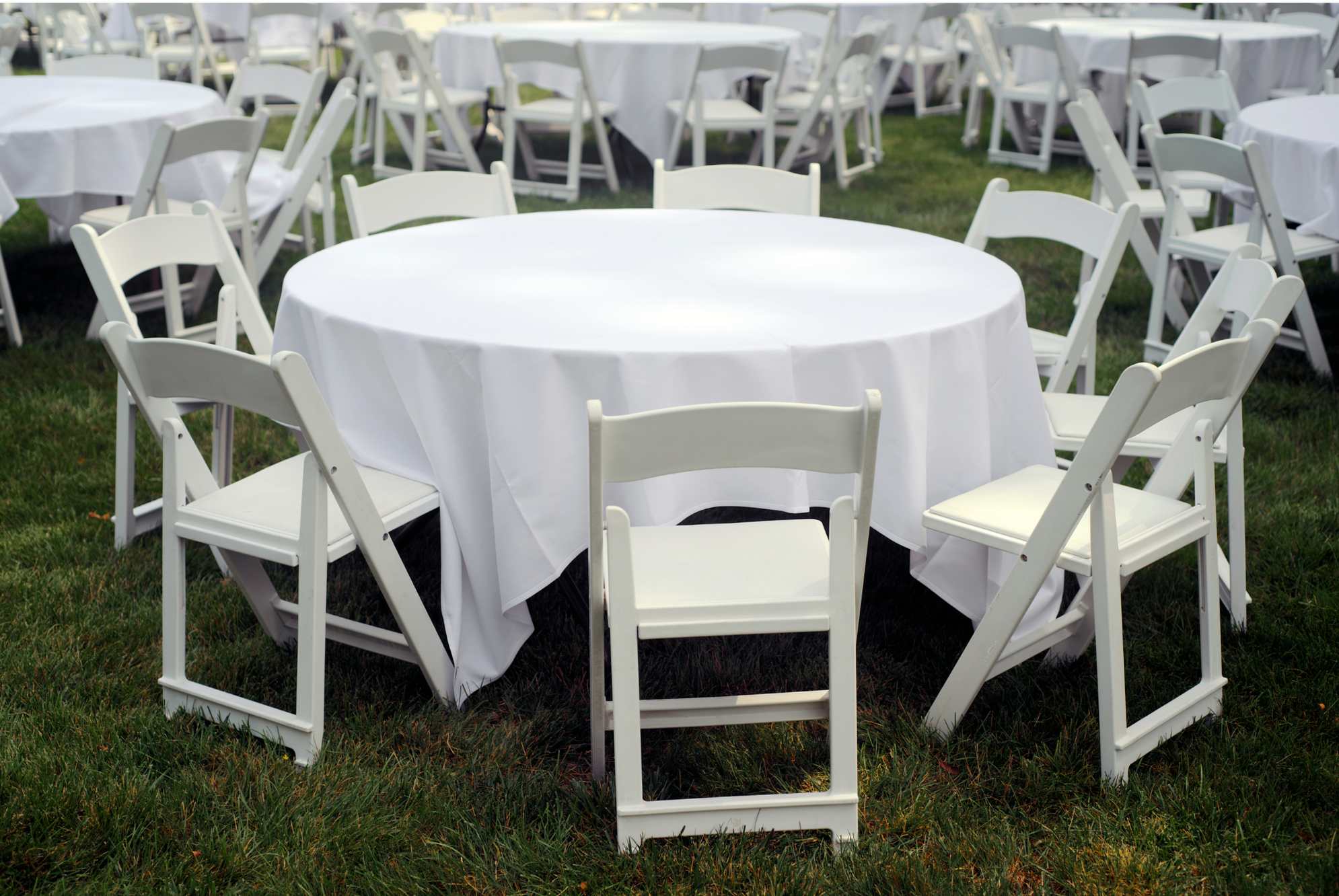 Round table with white tablecloth and white folding chairs set up outdoors on grass.