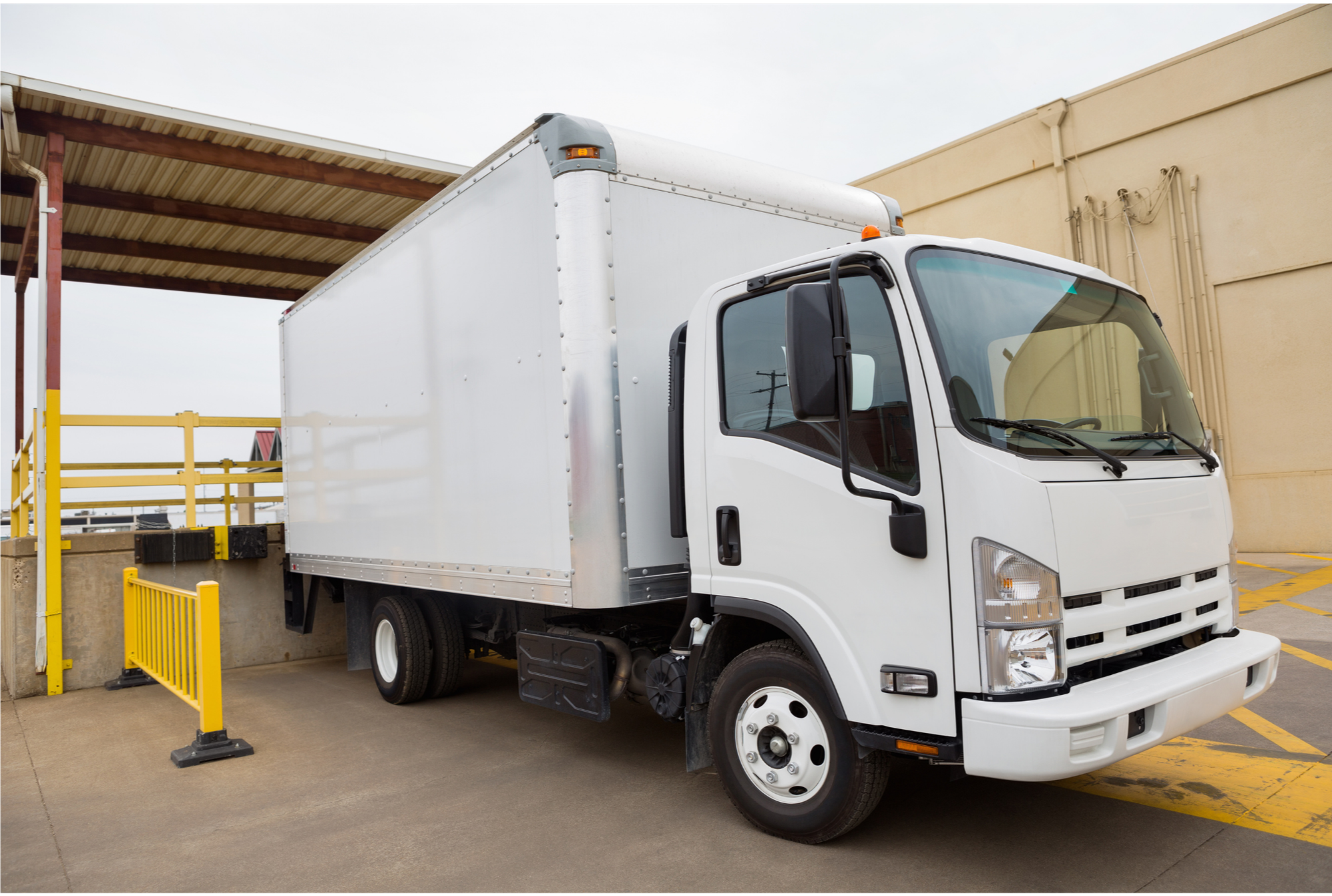 White delivery truck parked at a loading dock with yellow safety barriers in an industrial area.