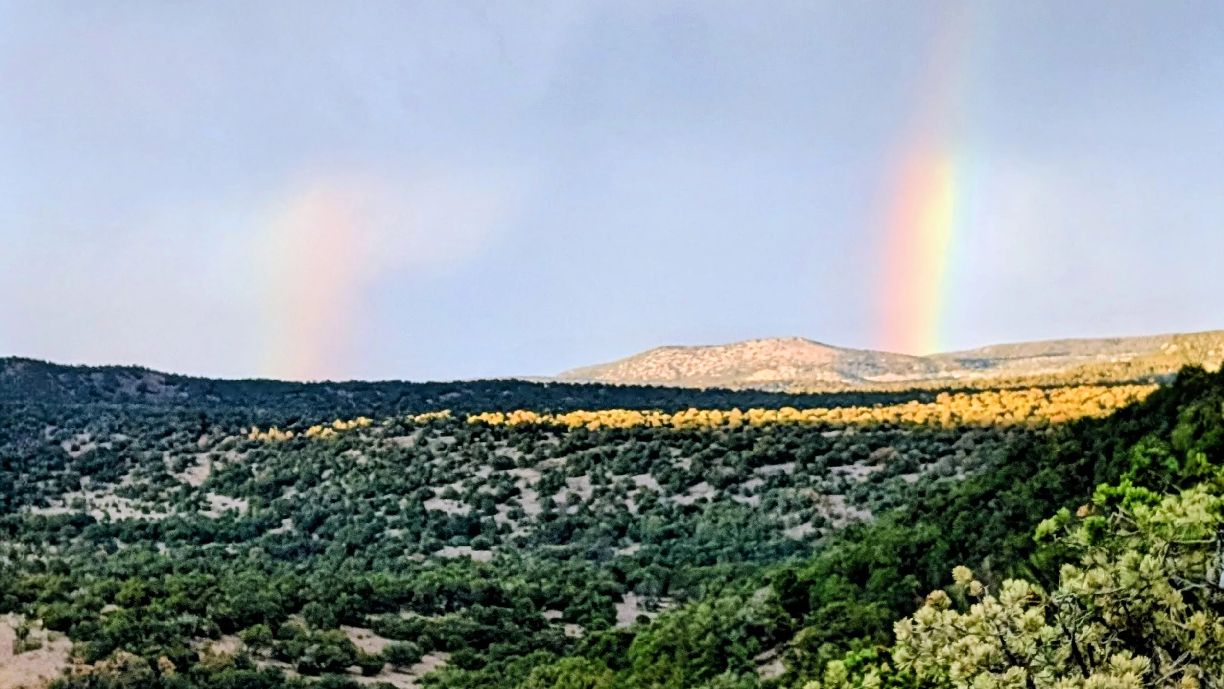 A landscape view of rolling hills with green and yellow trees, a mountain in the background, and two faint rainbows in the sky above.