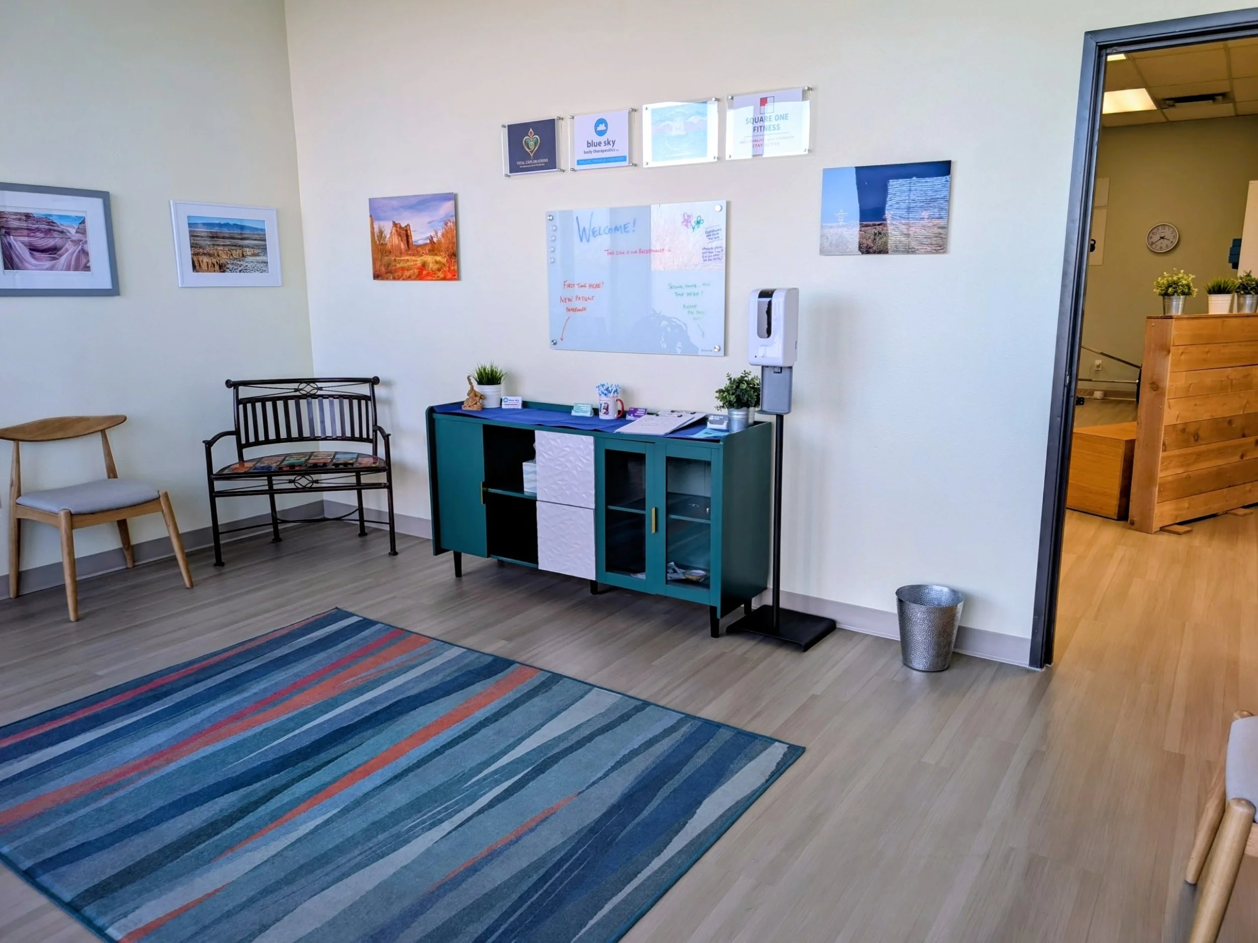 Waiting area with two chairs, a colorful rug, walls decorated with landscape photographs, a whiteboard with a welcome message, a teal cabinet with potted plants, and a hand sanitizer dispenser on a stand.