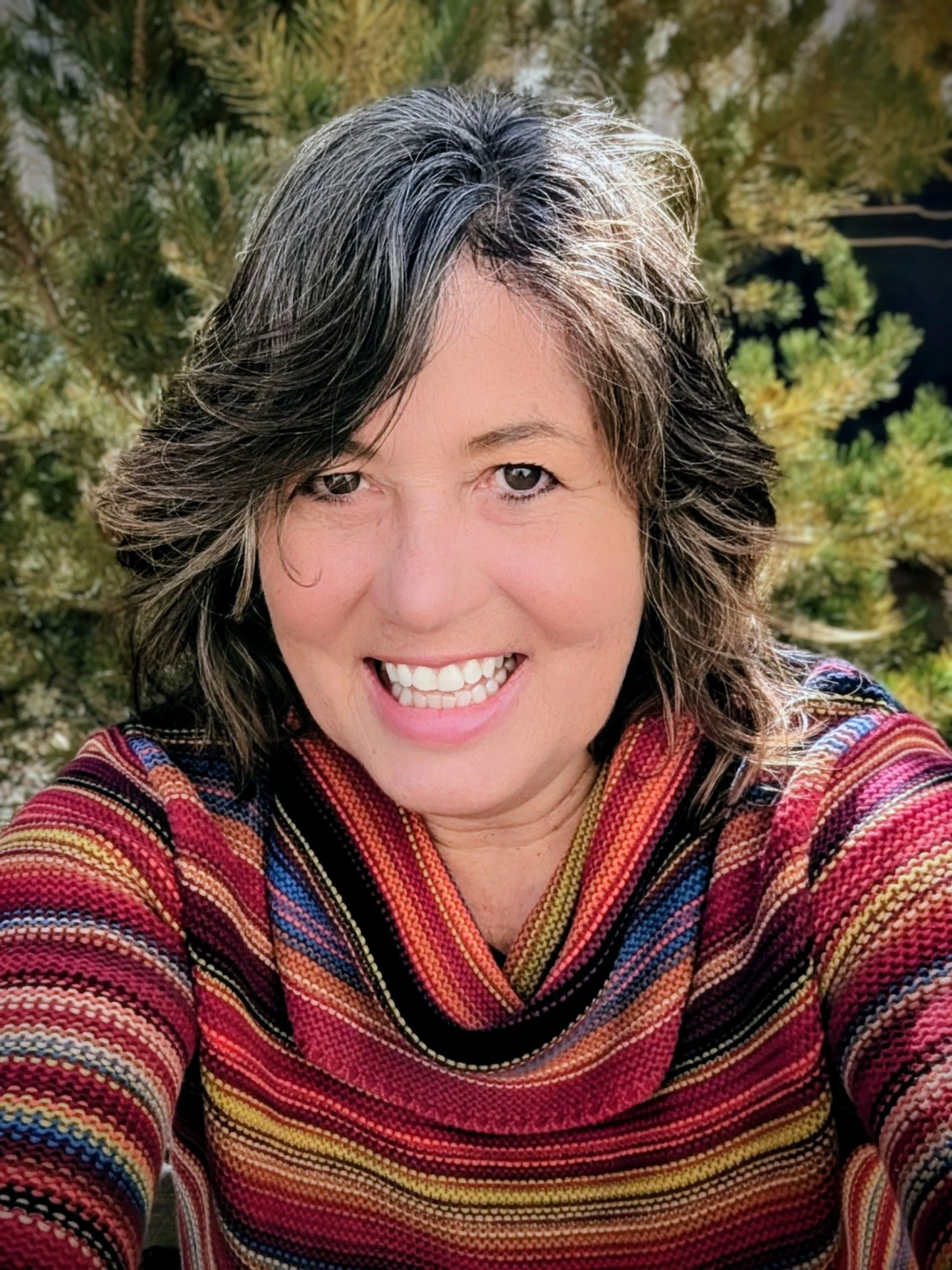 A woman with short, wavy gray hair smiling outdoors, wearing a colorful striped sweater, with green foliage in the background.
