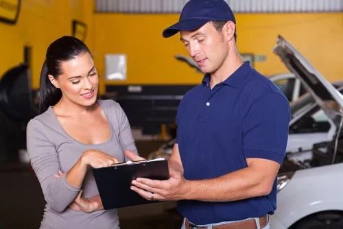 A woman and a man discussing in a garage, with a car and tools in the background.