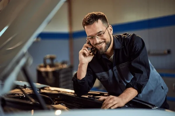 A man with glasses and a beard smiling while talking on a phone, working on a car in a garage.