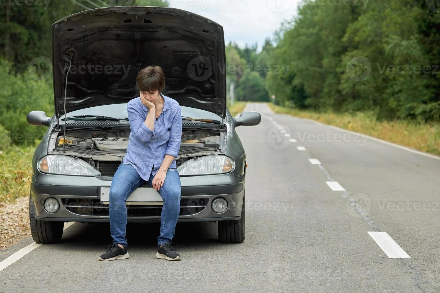 A woman sitting on the front bumper of a car with its hood open on a deserted road surrounded by trees, looking worried and holding her head in distress.