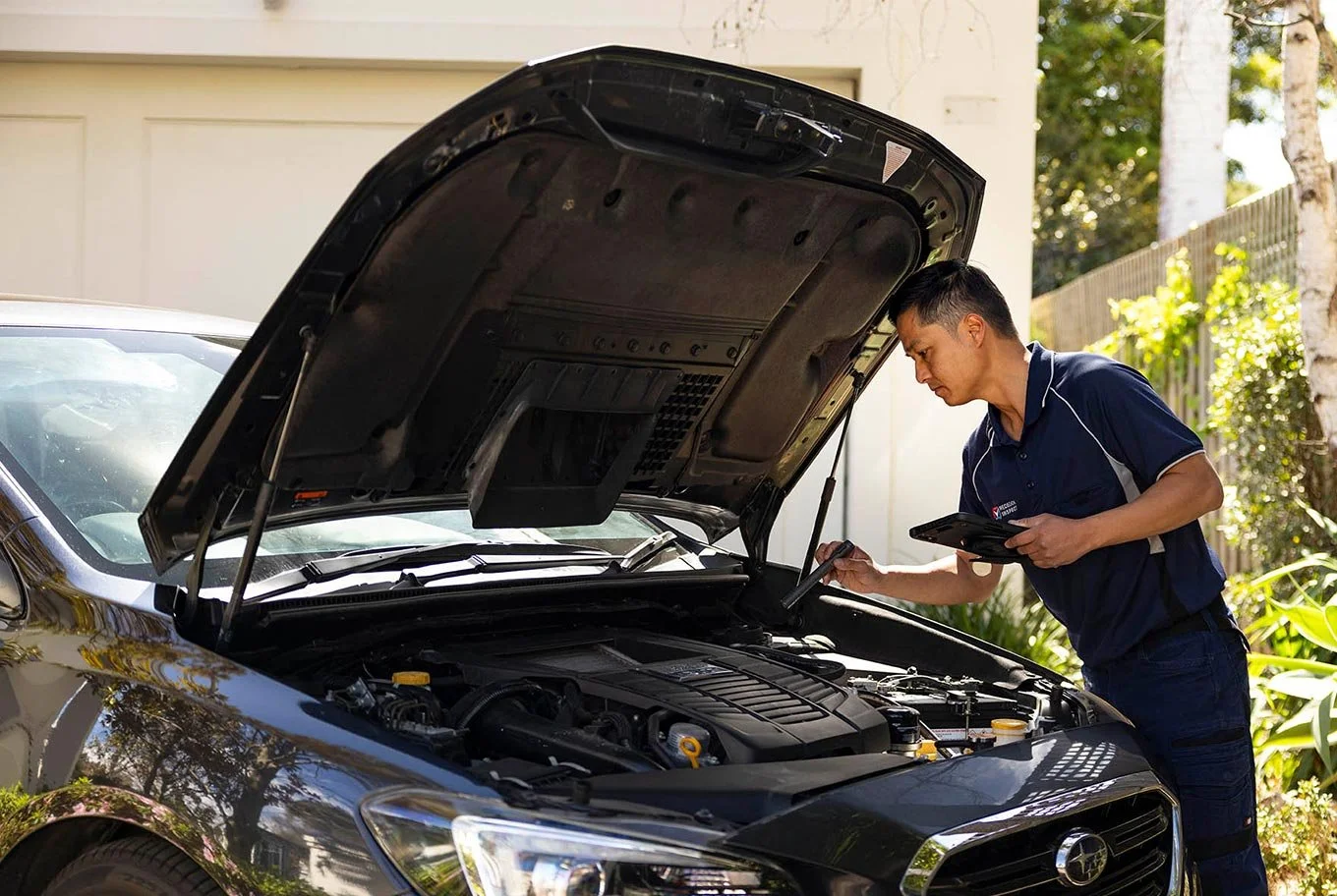 A mechanic inspecting a car engine with a tablet outside in a driveway.
