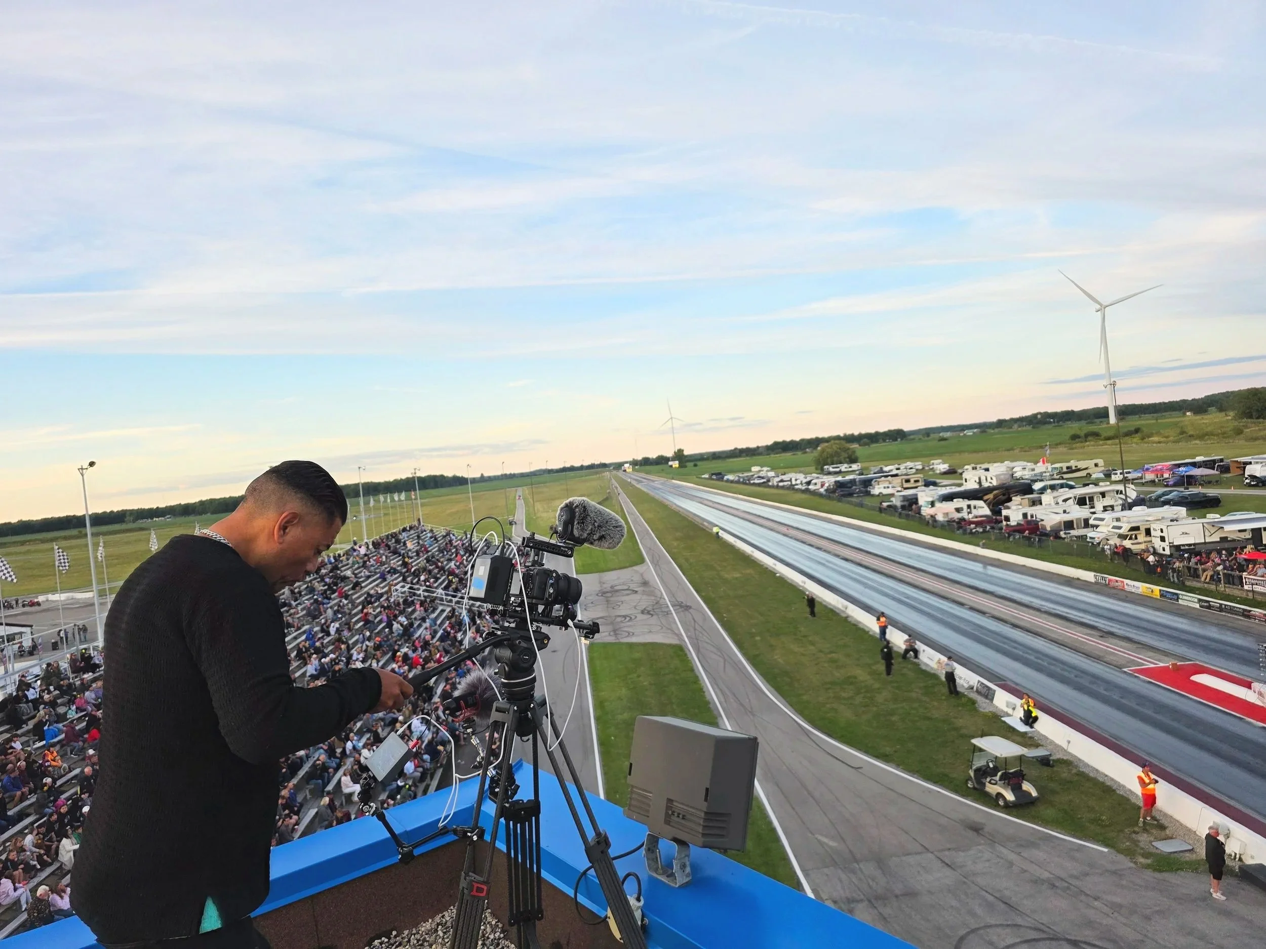 Live footage being recorded at a drag racing event with a camera operator on a tower, a race track with cars, and a large crowd in the stands, with parked trailers and wind turbines in the background.