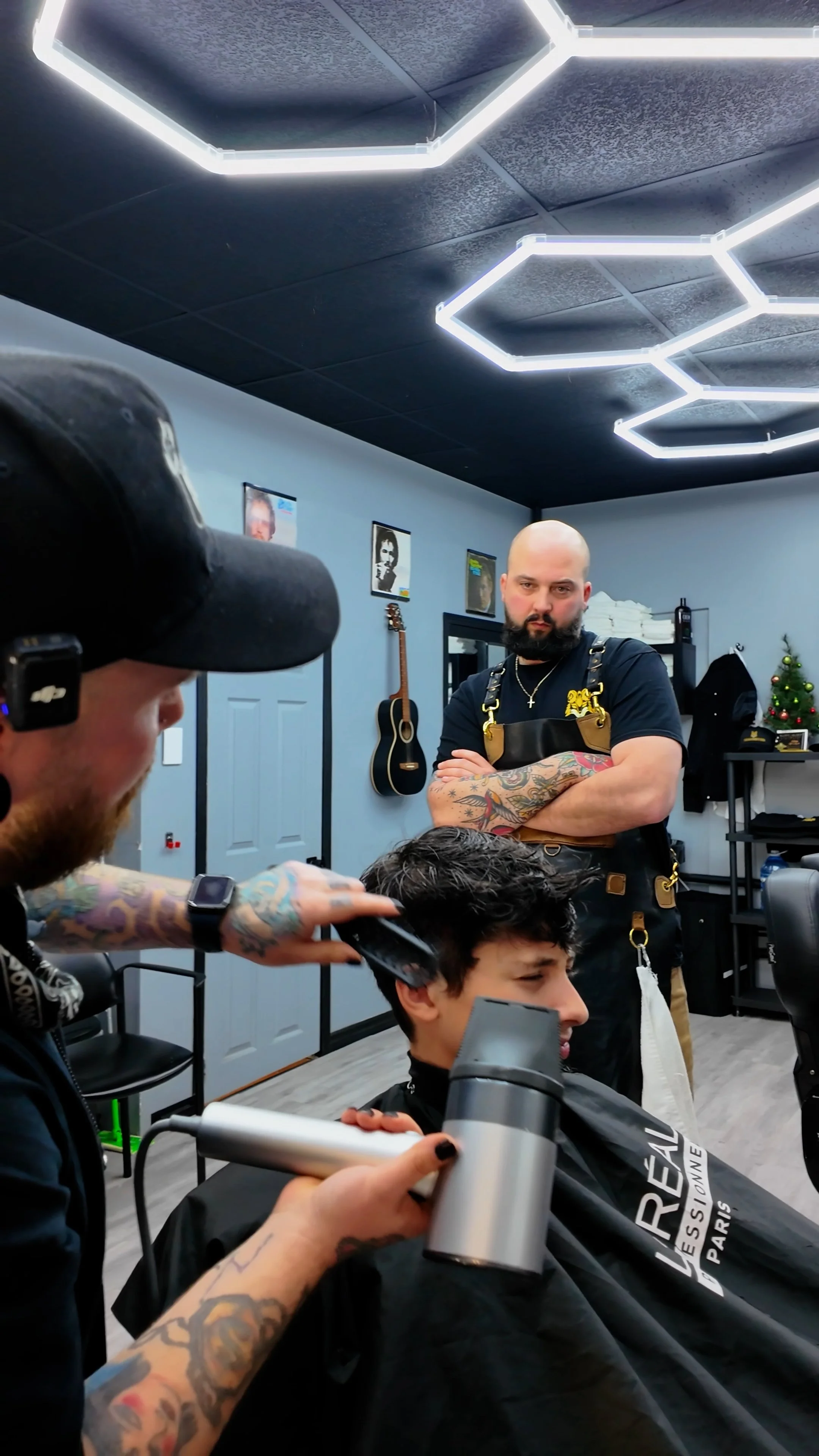 A man getting a haircut in a modern barber shop with hexagon-shaped lights on the ceiling. The barber is holding hair clippers and a spray bottle, while the customer is seated with a black cape. There are framed pictures, a guitar, a small Christmas tree, and shelves in the background