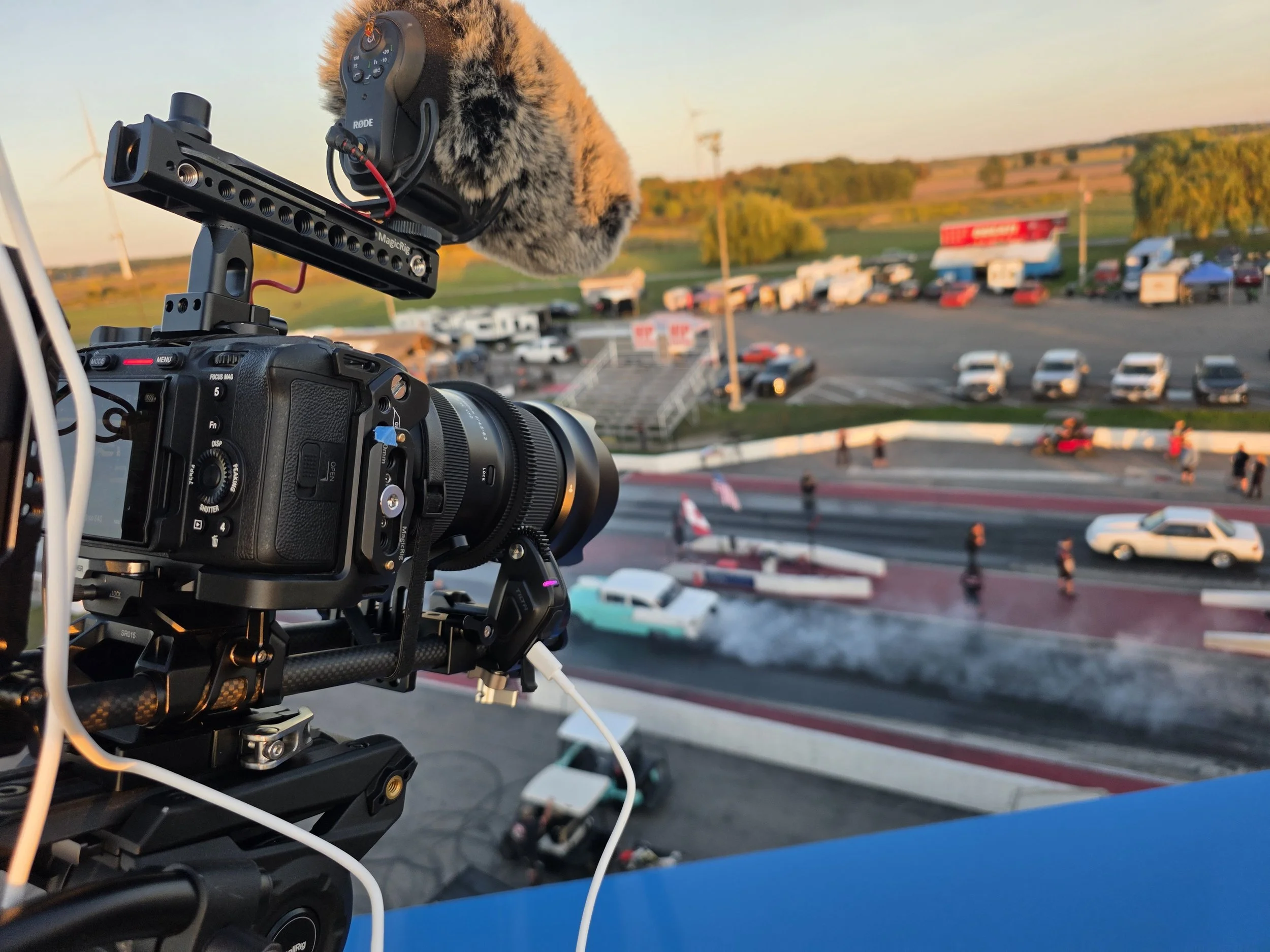 A professional camera with a microphone mounted on top filming a drag race at a racetrack during sunset, with cars and spectators in the background.