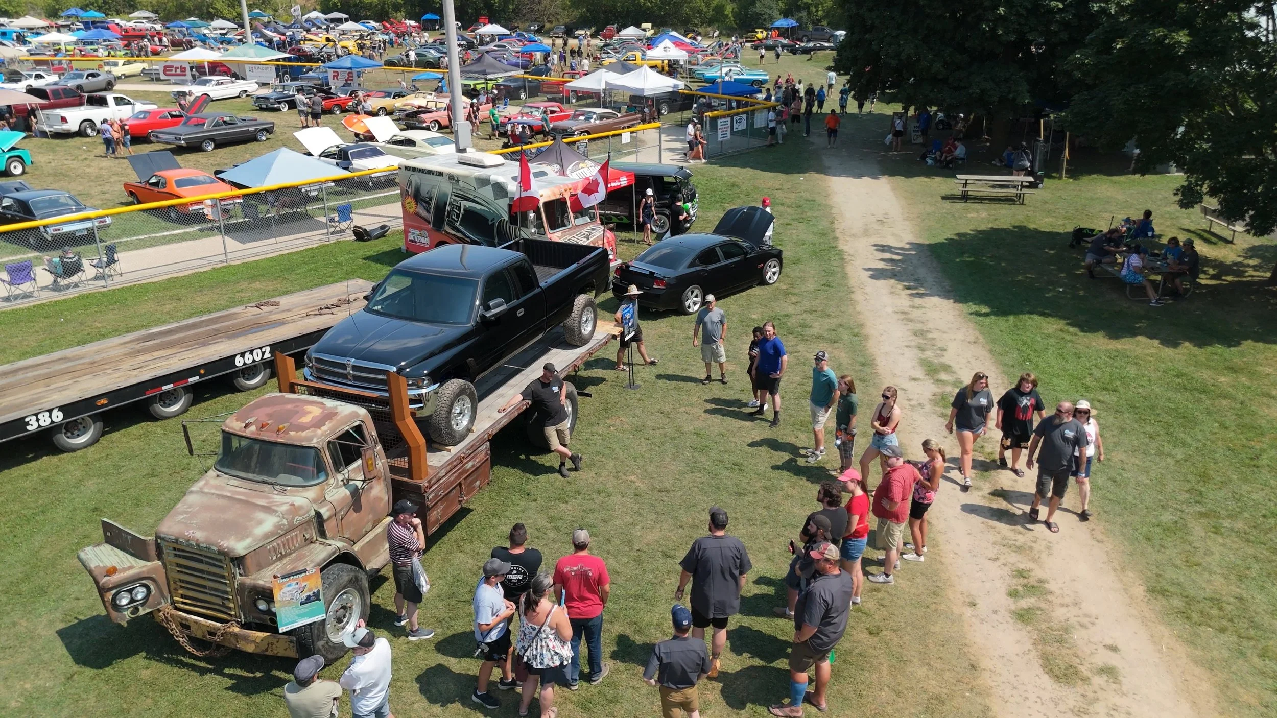Many classic cars on display at a car show with a line of people waiting to see them, a truck with a rusted truck hood, black pickup truck, and many other vintage cars parked in the background.