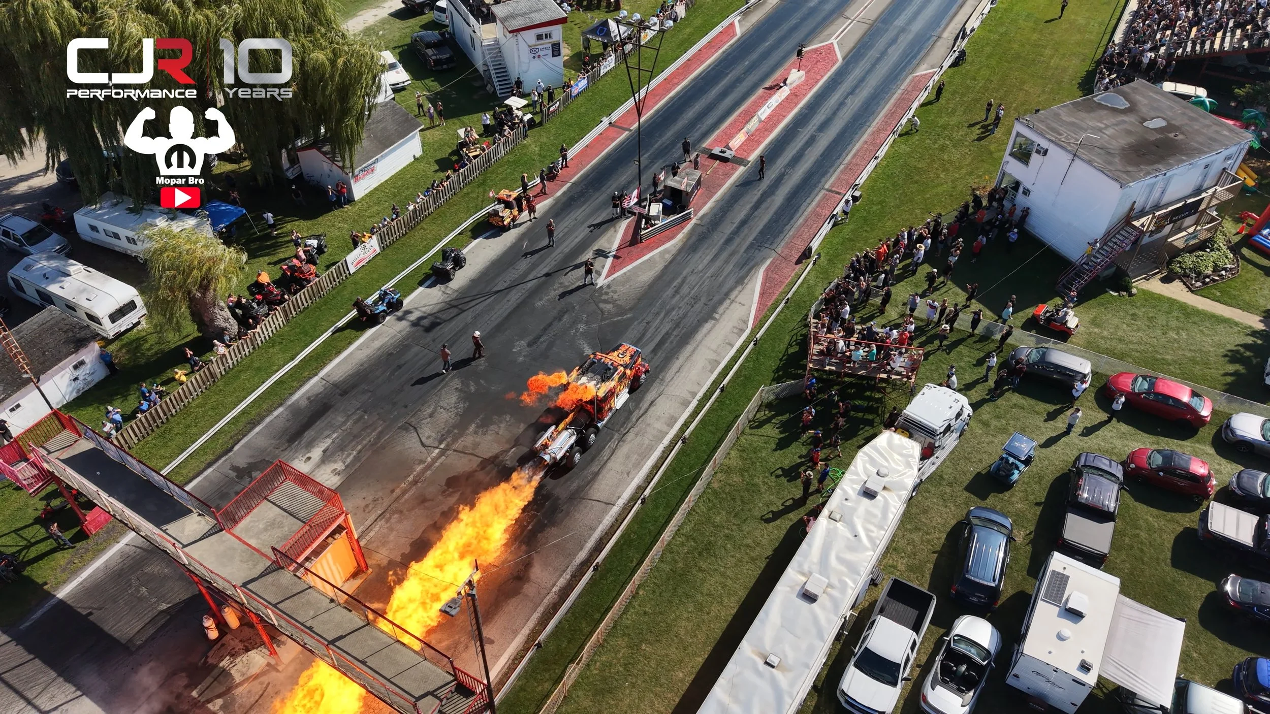 An aerial view of a drag racing event with a car on fire at the starting line, spectators on both sides, and several vehicles parked nearby.