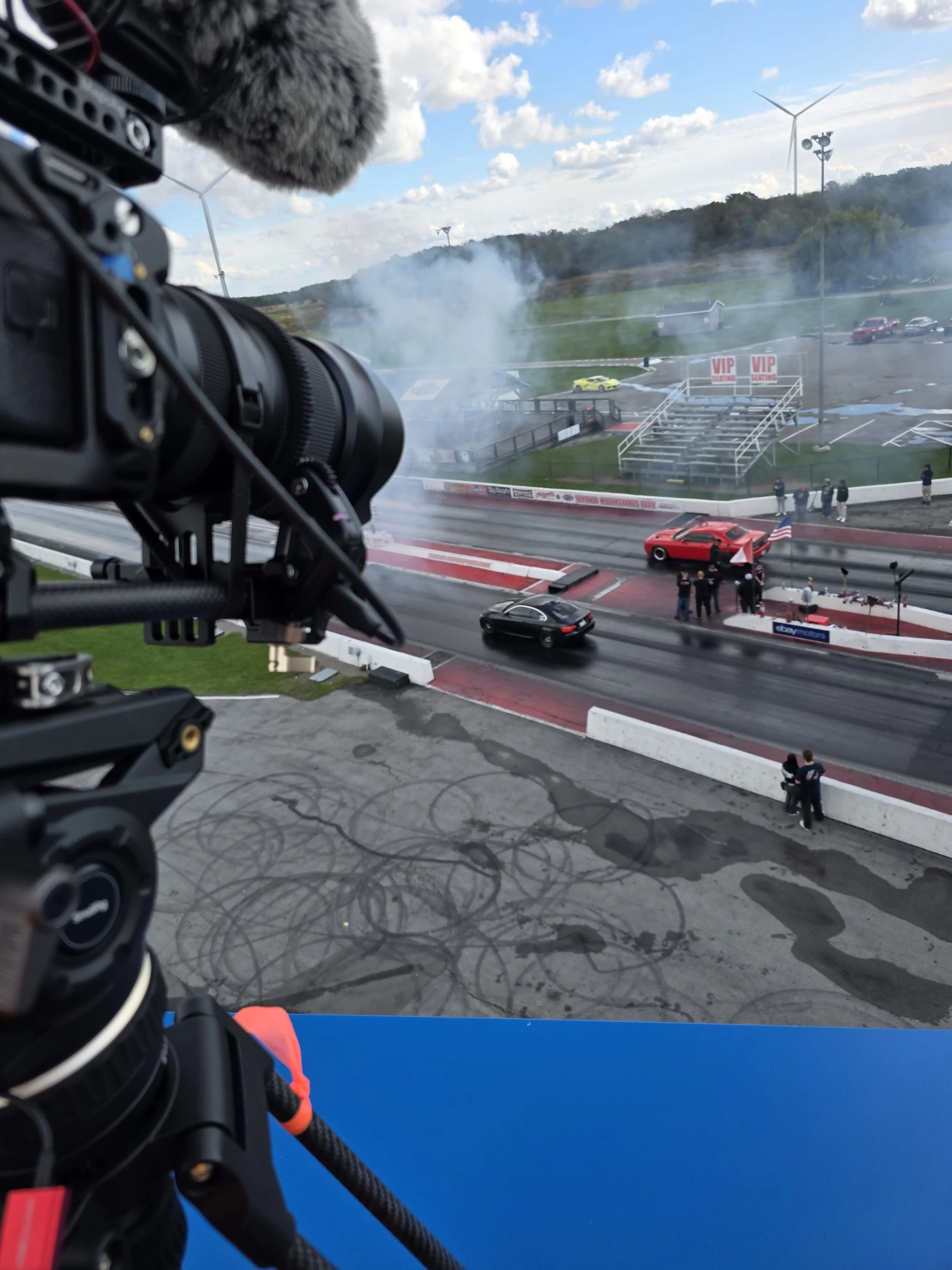 Race track with cars, smoke, and spectators. Camera setup in foreground capturing the track, finishes line, and a car with an American flag. Wind turbines in background.